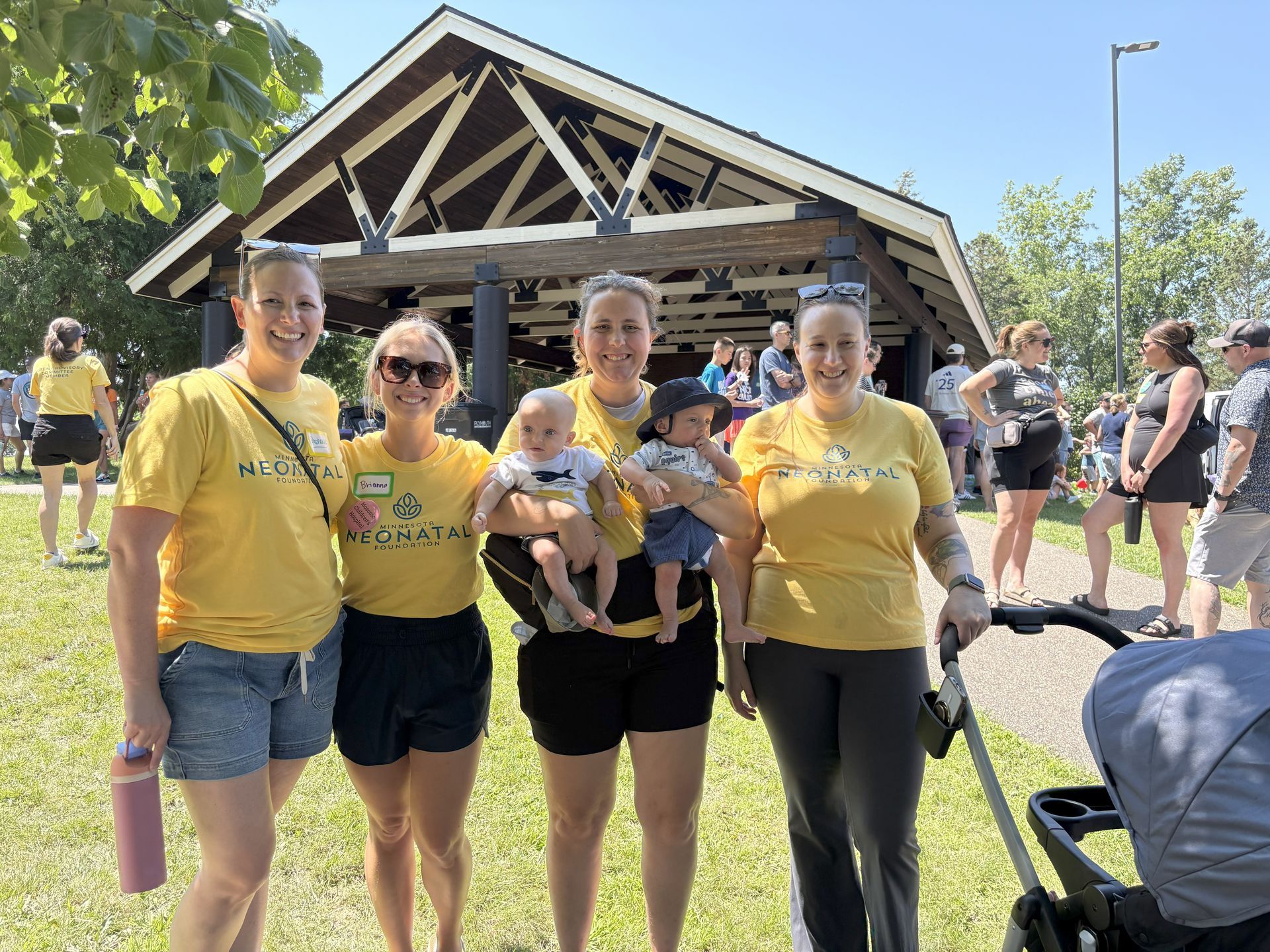 Group of women in yellow shirts, holding babies, by a park pavilion on a sunny day.