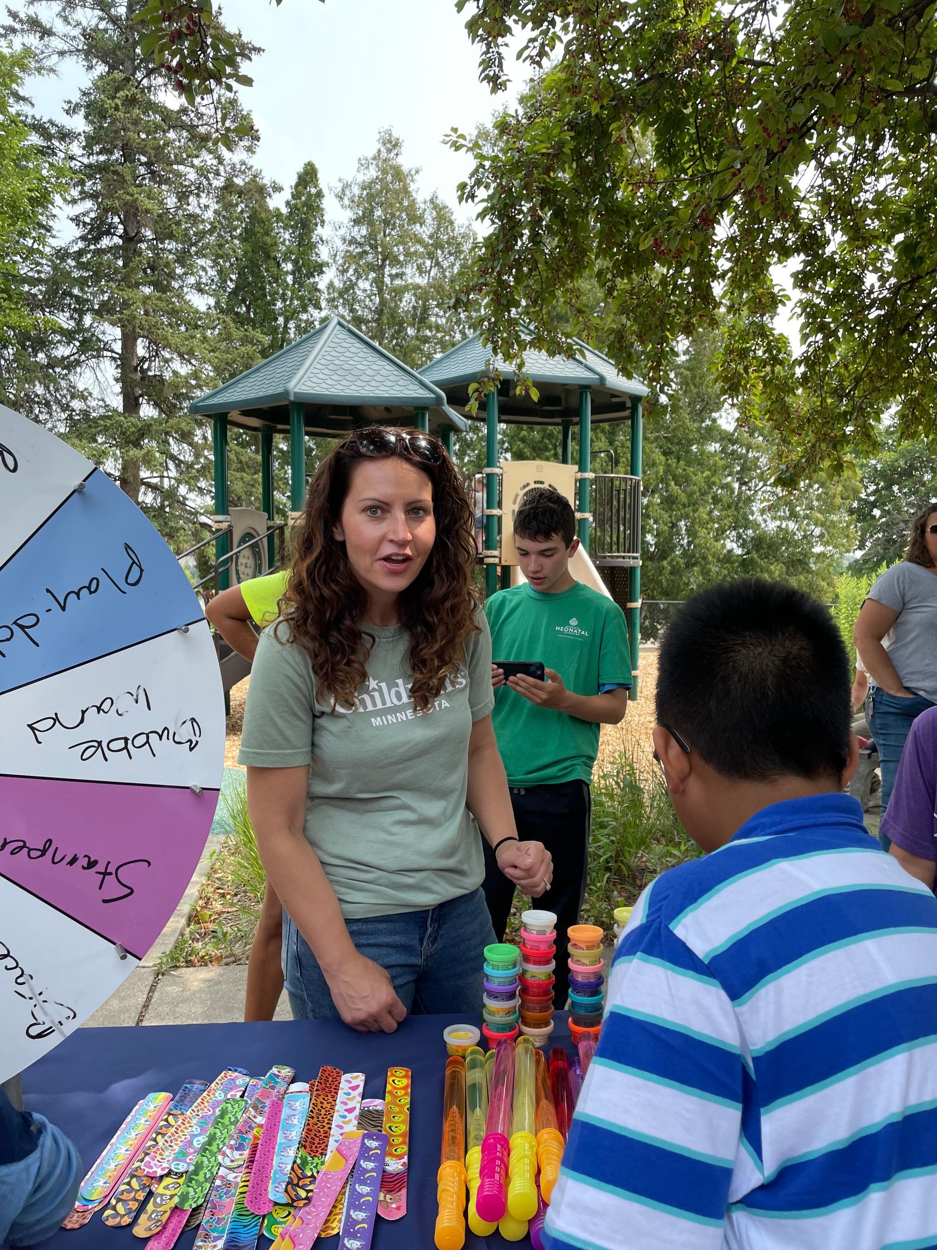 Woman at a game booth with prizes, offering to a child. A spinning wheel and playground are in the background.
