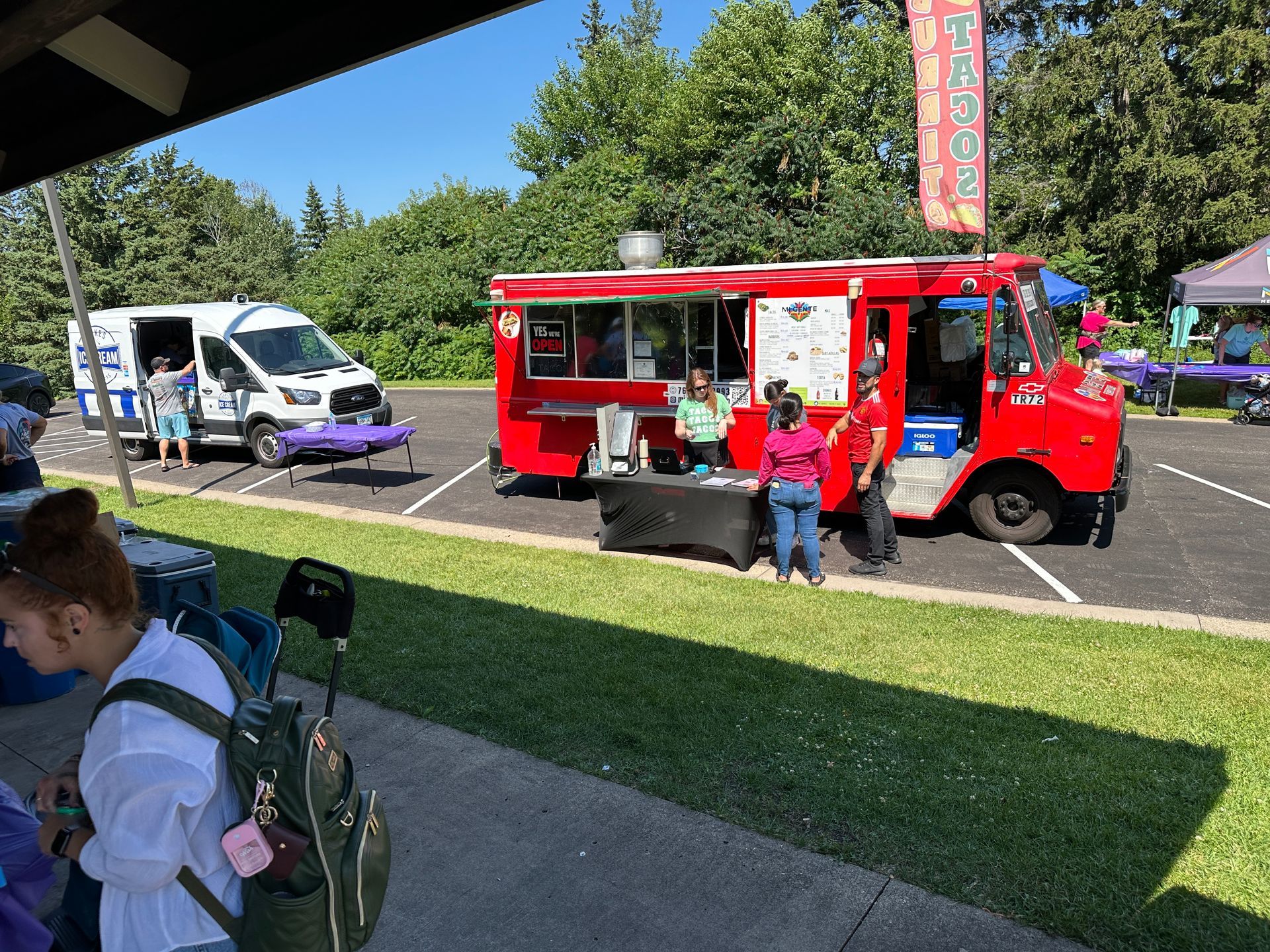Red food truck in parking lot, people ordering food. White van and green grass visible.