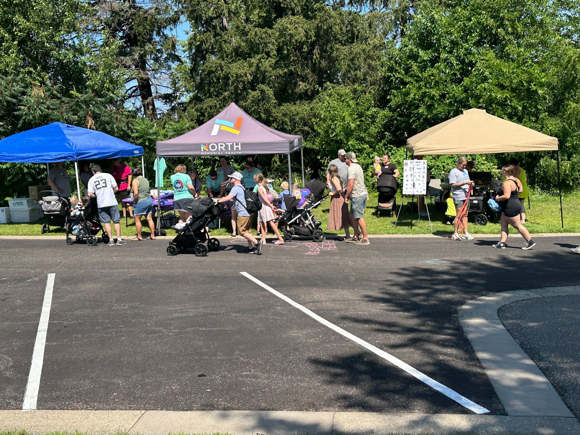 Outdoor market with tents, people, and strollers. People browsing and walking near booths in a parking lot.
