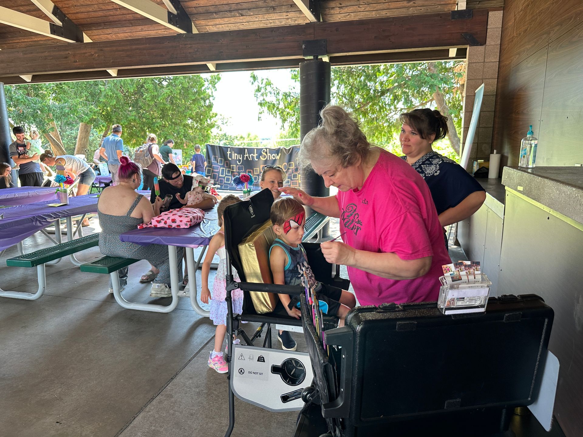 A woman adjusts a child's hair in a wheelchair at an outdoor gathering with tables and people.