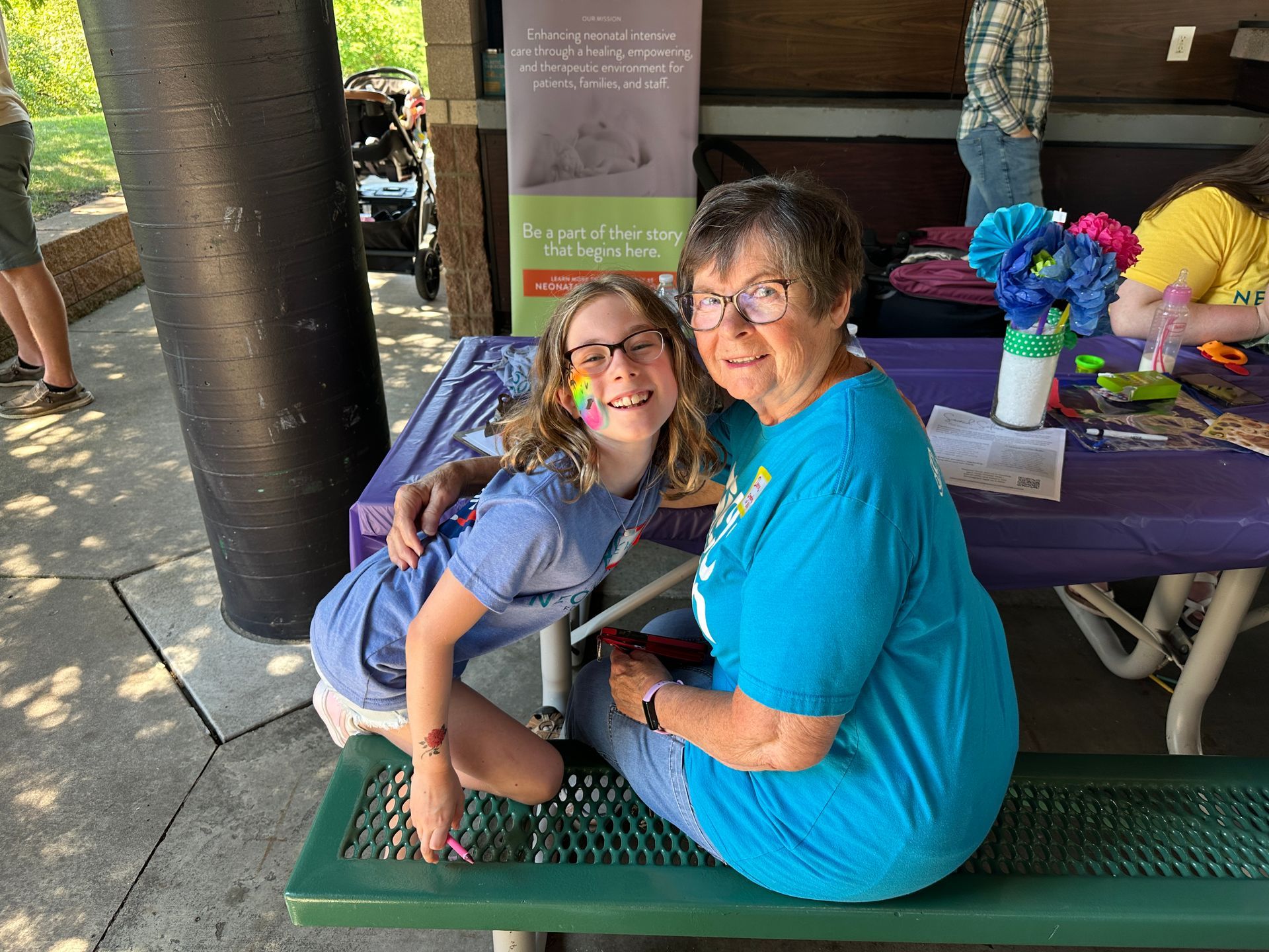 Girl with face paint hugs woman at a purple table outdoors. Both smile, next to a sign.