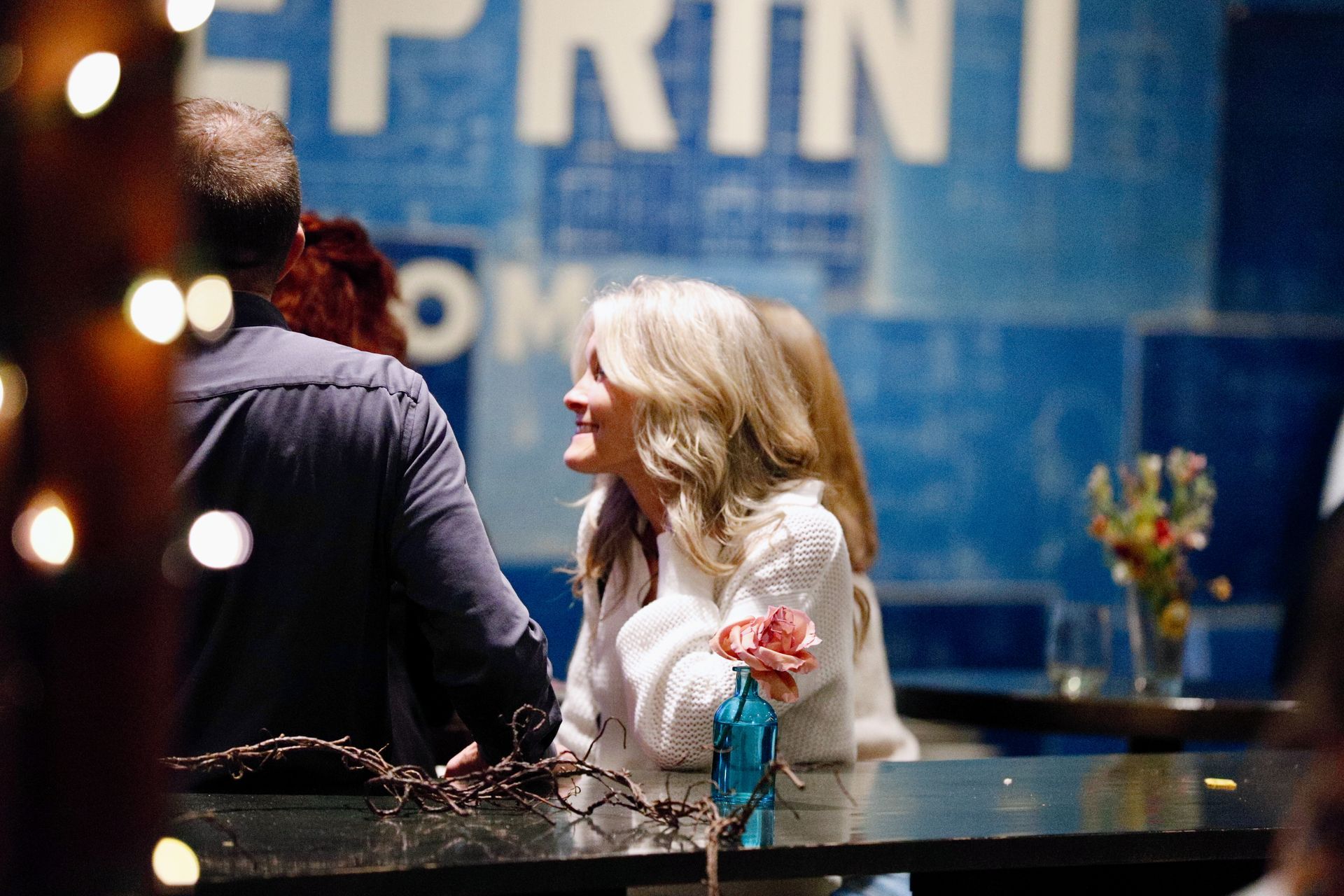 Woman smiles while standing near a bar, with other people and blue wall in background.