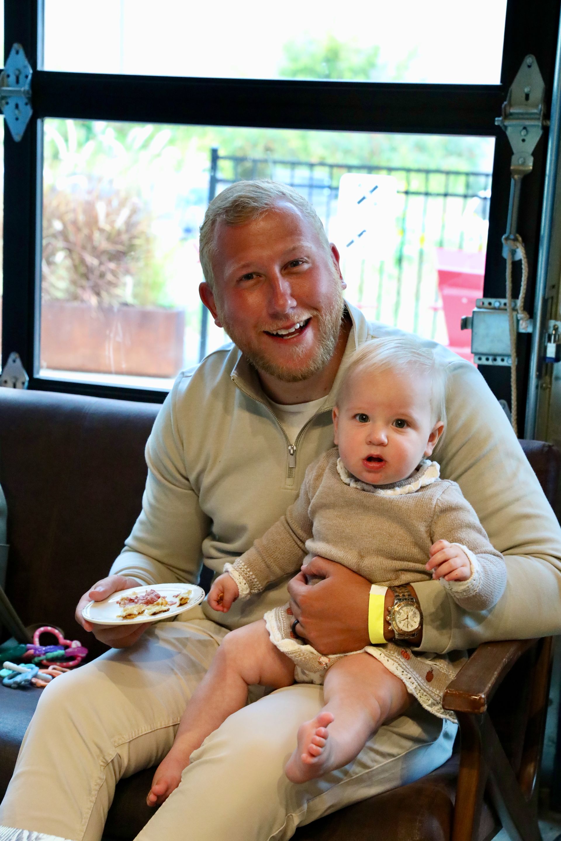 Man with blonde hair, smiling, holding baby wearing a sweater, seated indoors near a window.