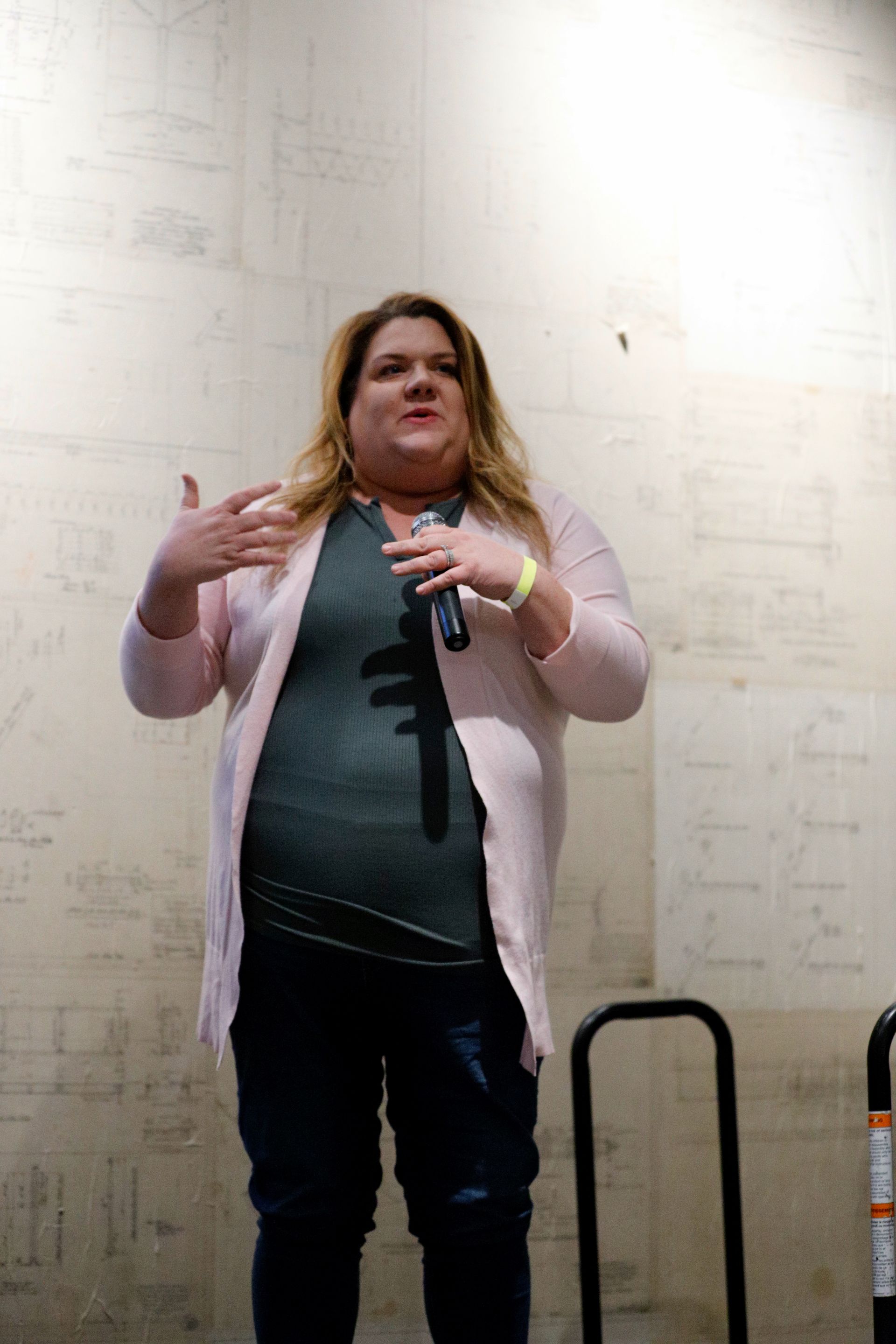 Woman speaking into microphone, gesturing with hands. Wearing pink cardigan, green top, jeans, standing in front of light wall.