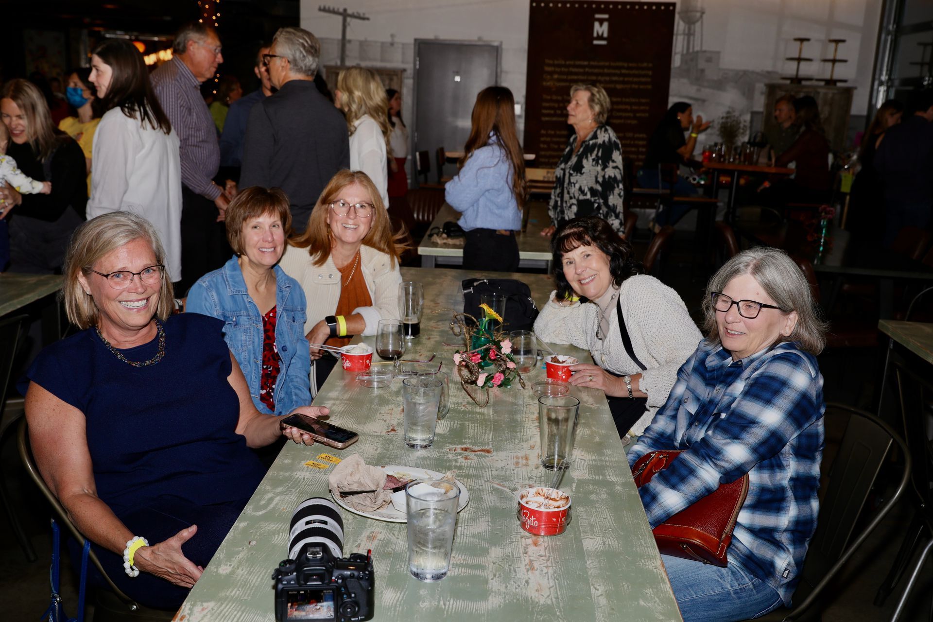 Group of women seated at a table, smiling at the camera, inside a dimly lit event space.
