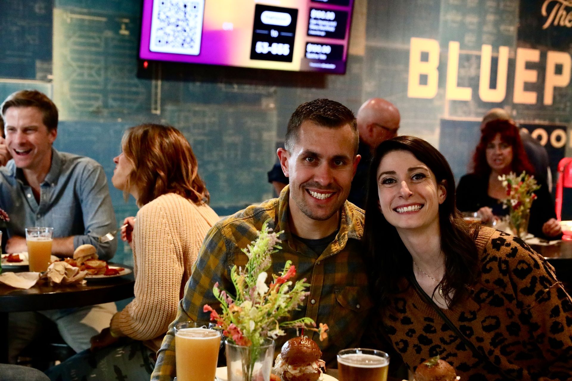 Couple smiles at camera in a bustling bar, surrounded by others. The Blue Print sign is visible.