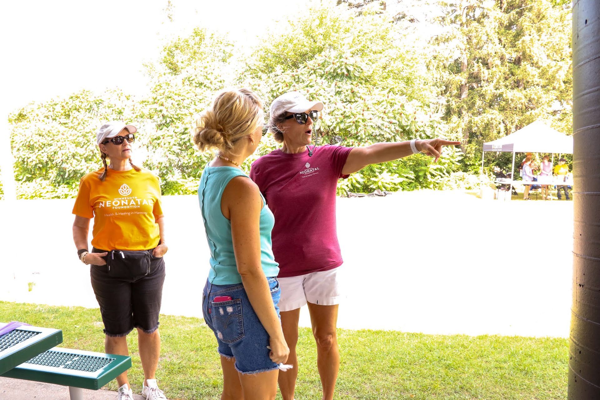 Three people outdoors; one points right, two wear hats; sunny day, park setting.
