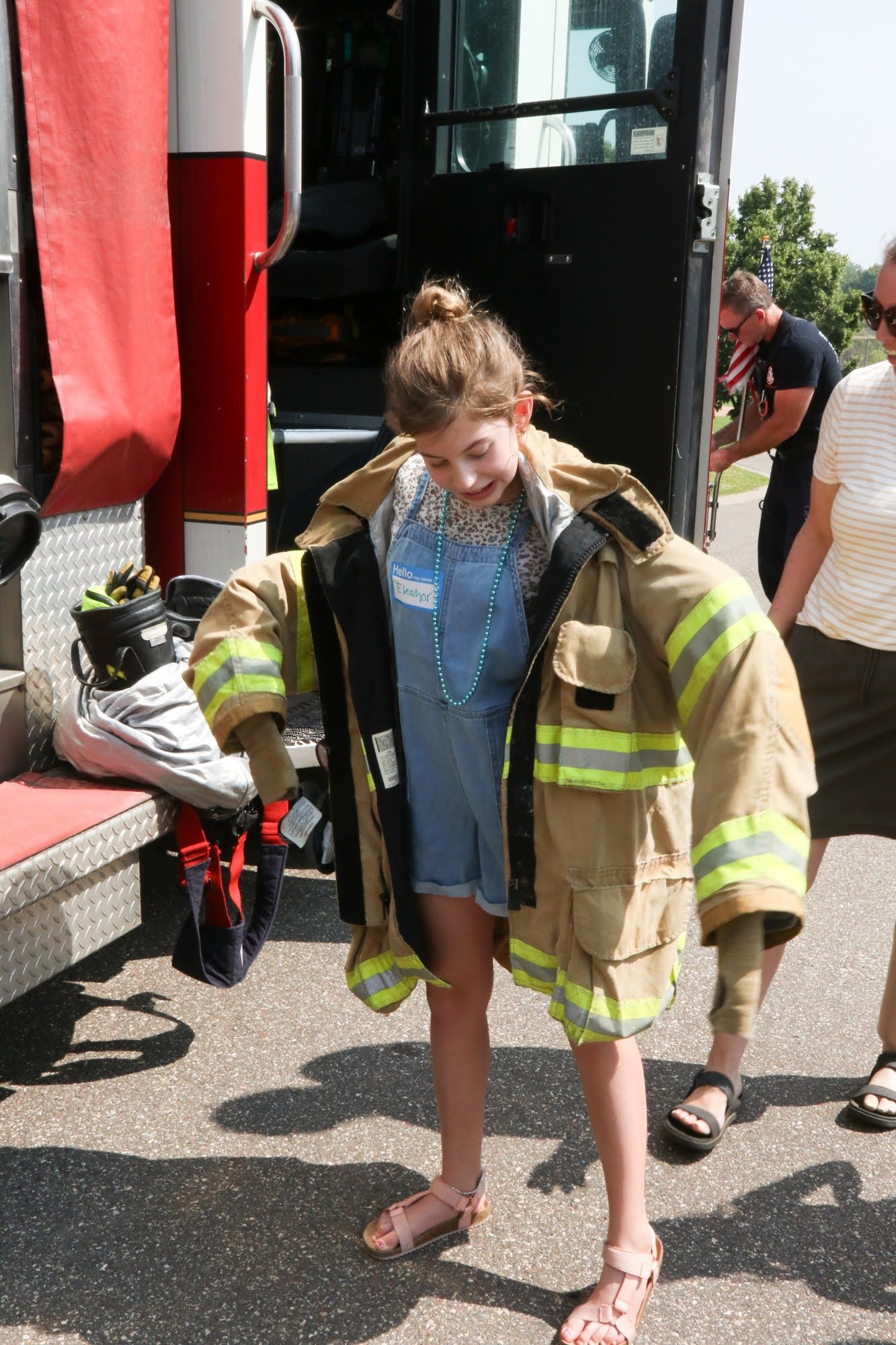 Girl in oversized firefighter jacket next to a fire truck; she's smiling.