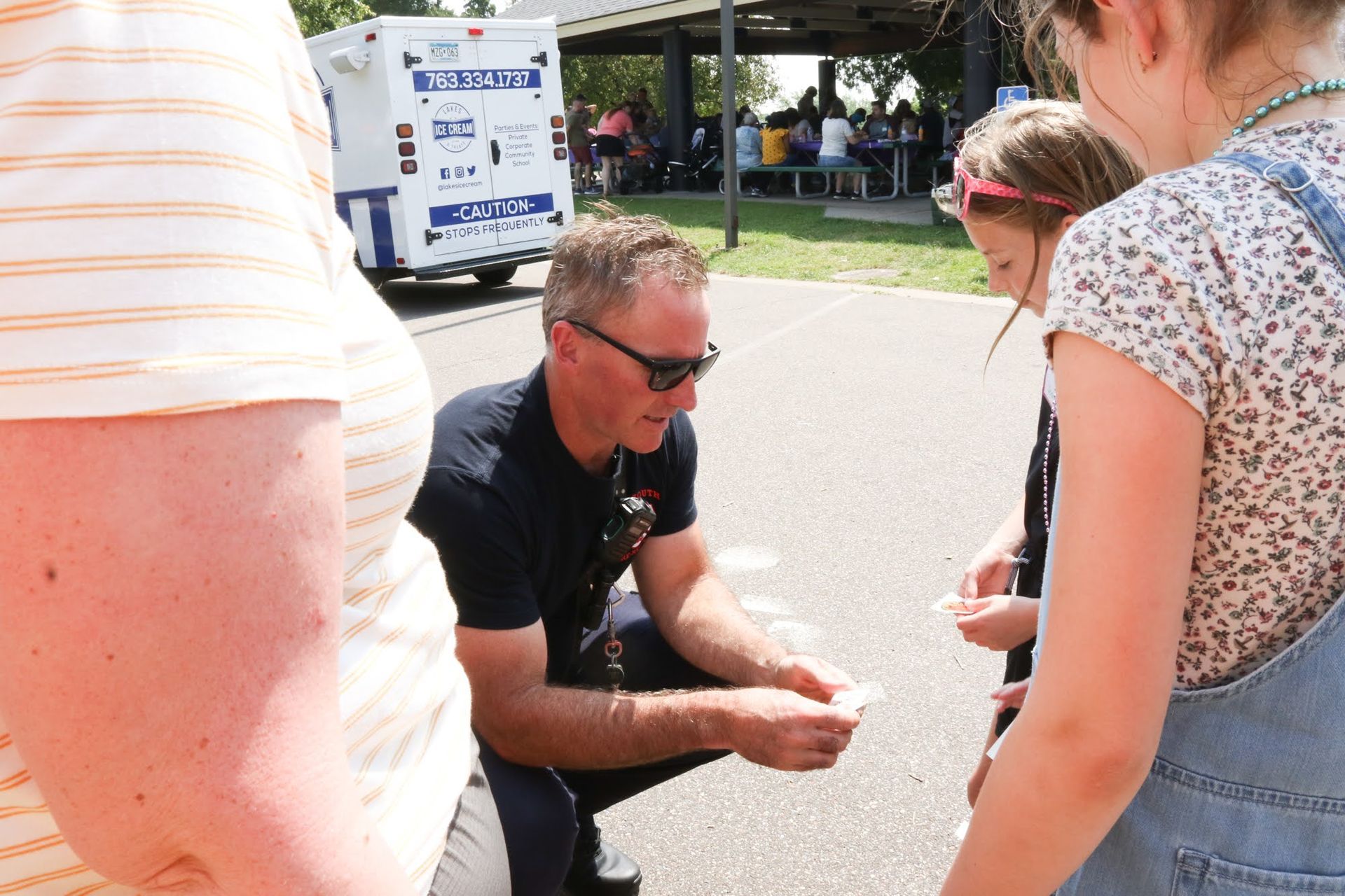 Man in sunglasses kneels, showing cards to two girls. An ice cream truck and people are in the background.