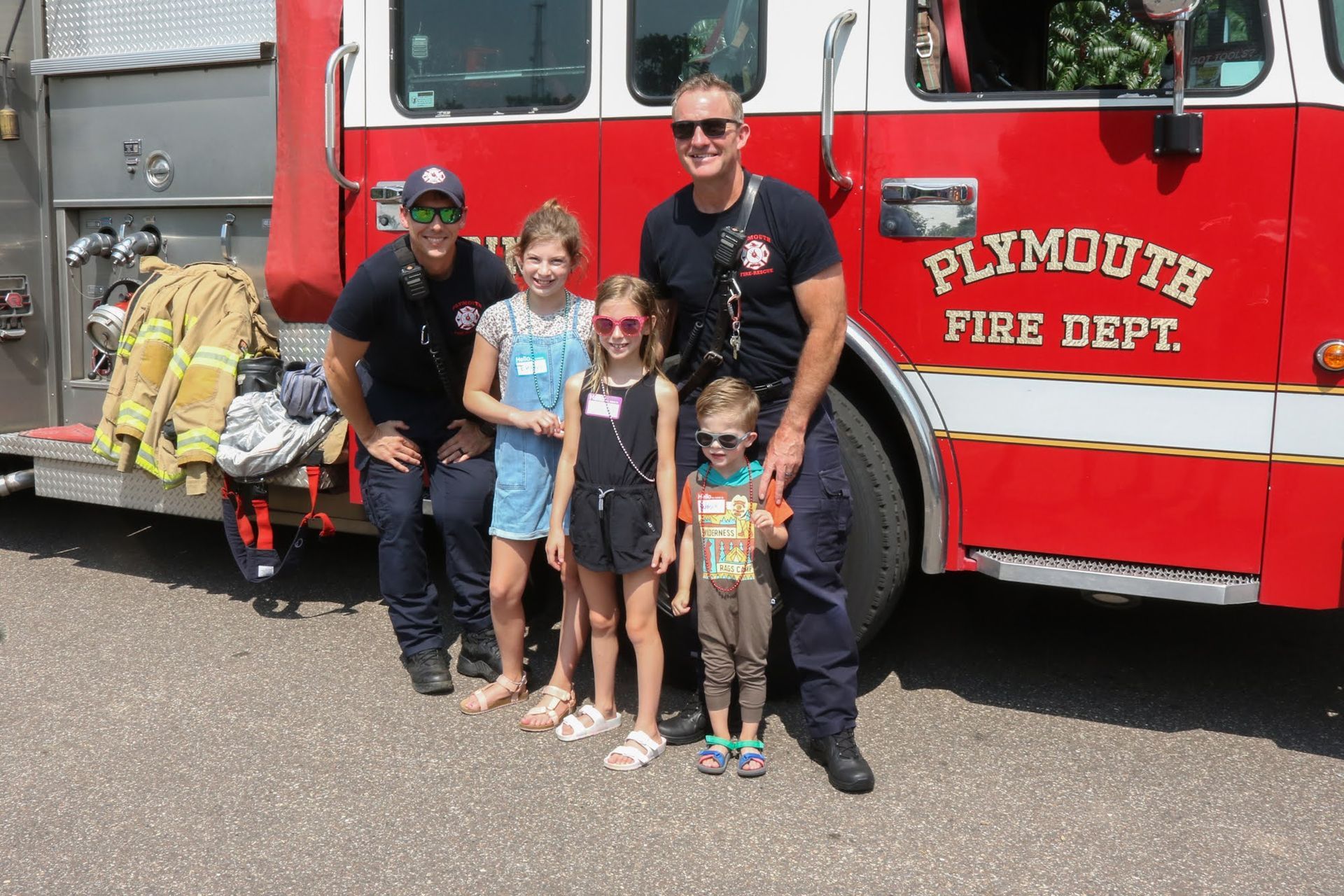 Two firefighters with four children in front of a red Plymouth Fire Dept. fire truck.