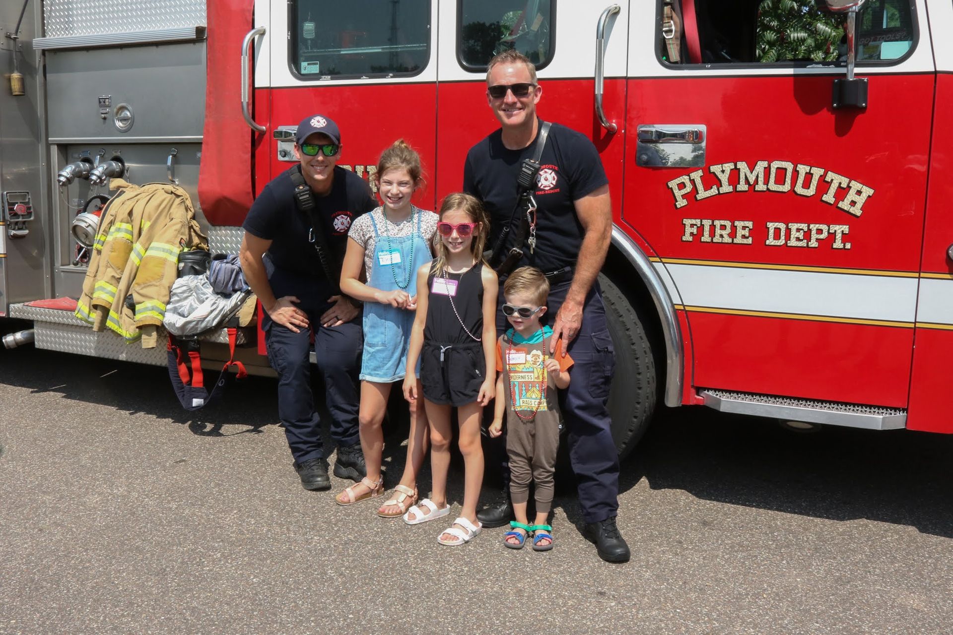 Two firefighters pose with three children next to a red fire truck that says Plymouth Fire Dept.