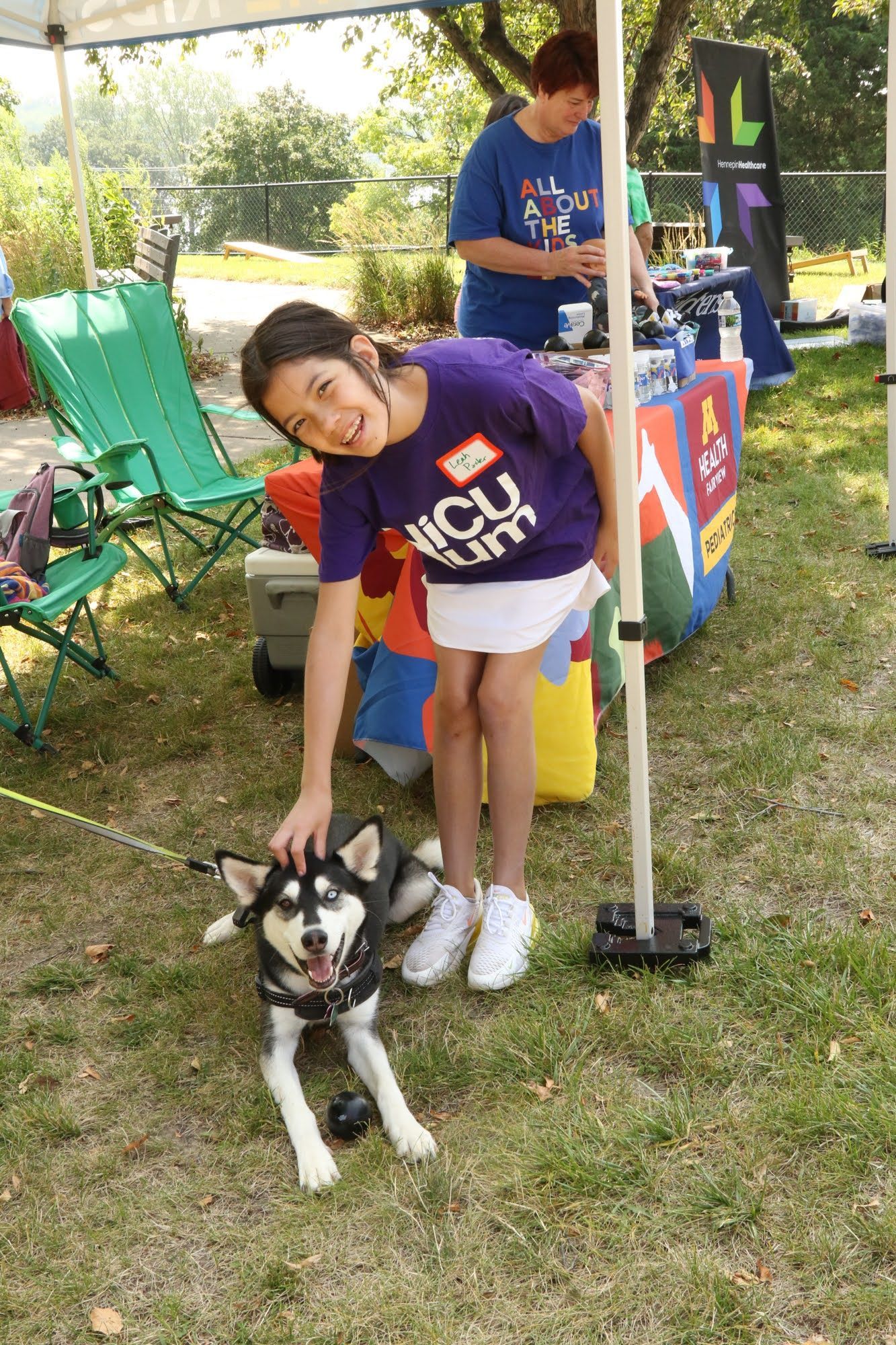 Girl petting a husky dog in a park, smiling. A woman in a blue shirt stands behind a table.