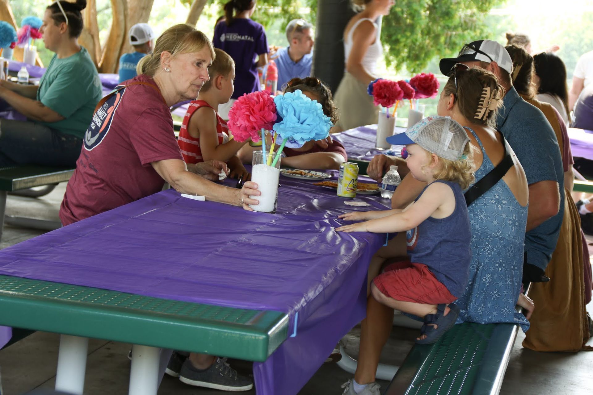 People seated at a picnic table with decorations. Woman serving drink to children. Outdoors, shaded area.