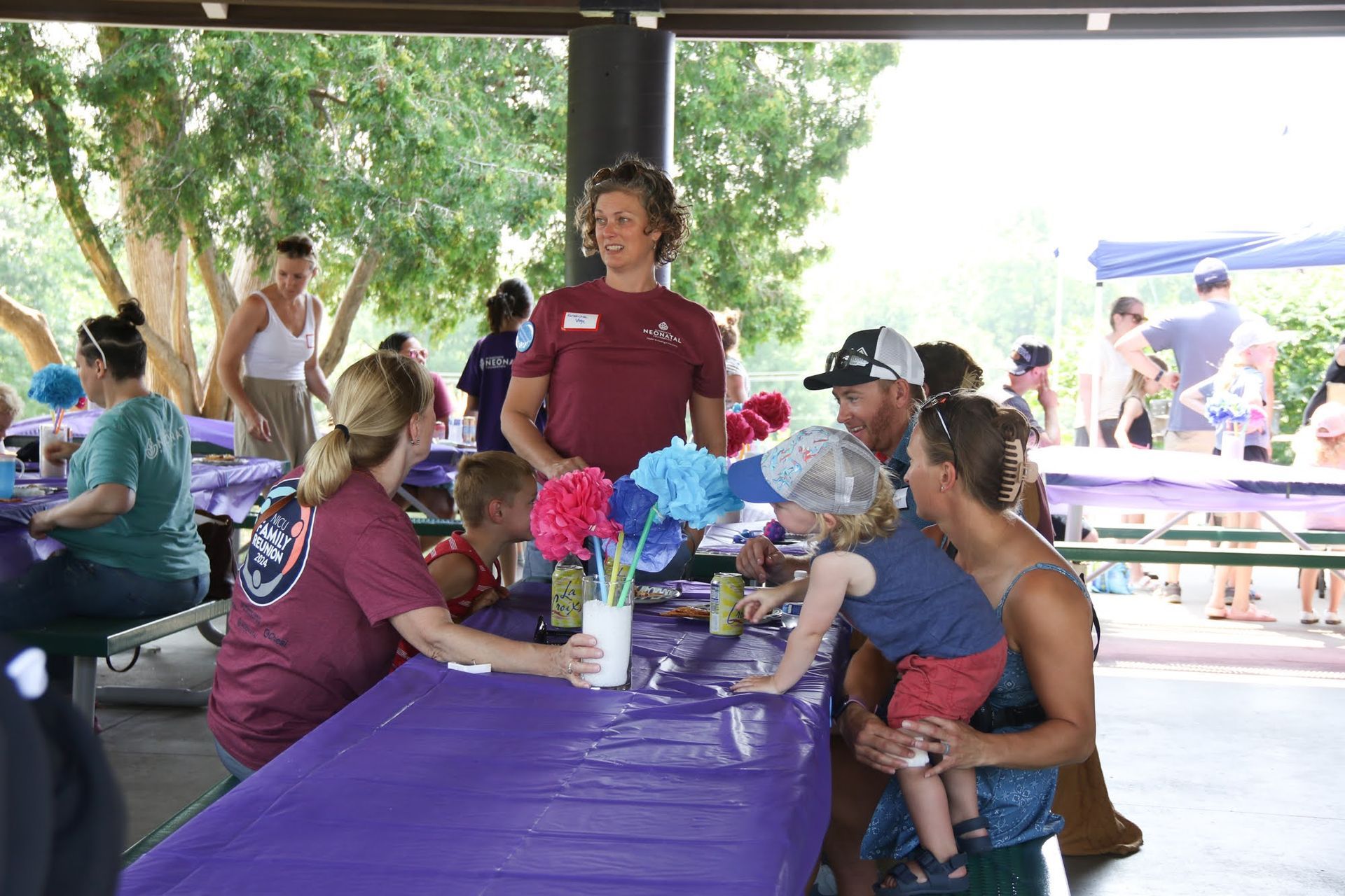 People at an outdoor picnic.  A person in a red shirt stands by a table with decorations.