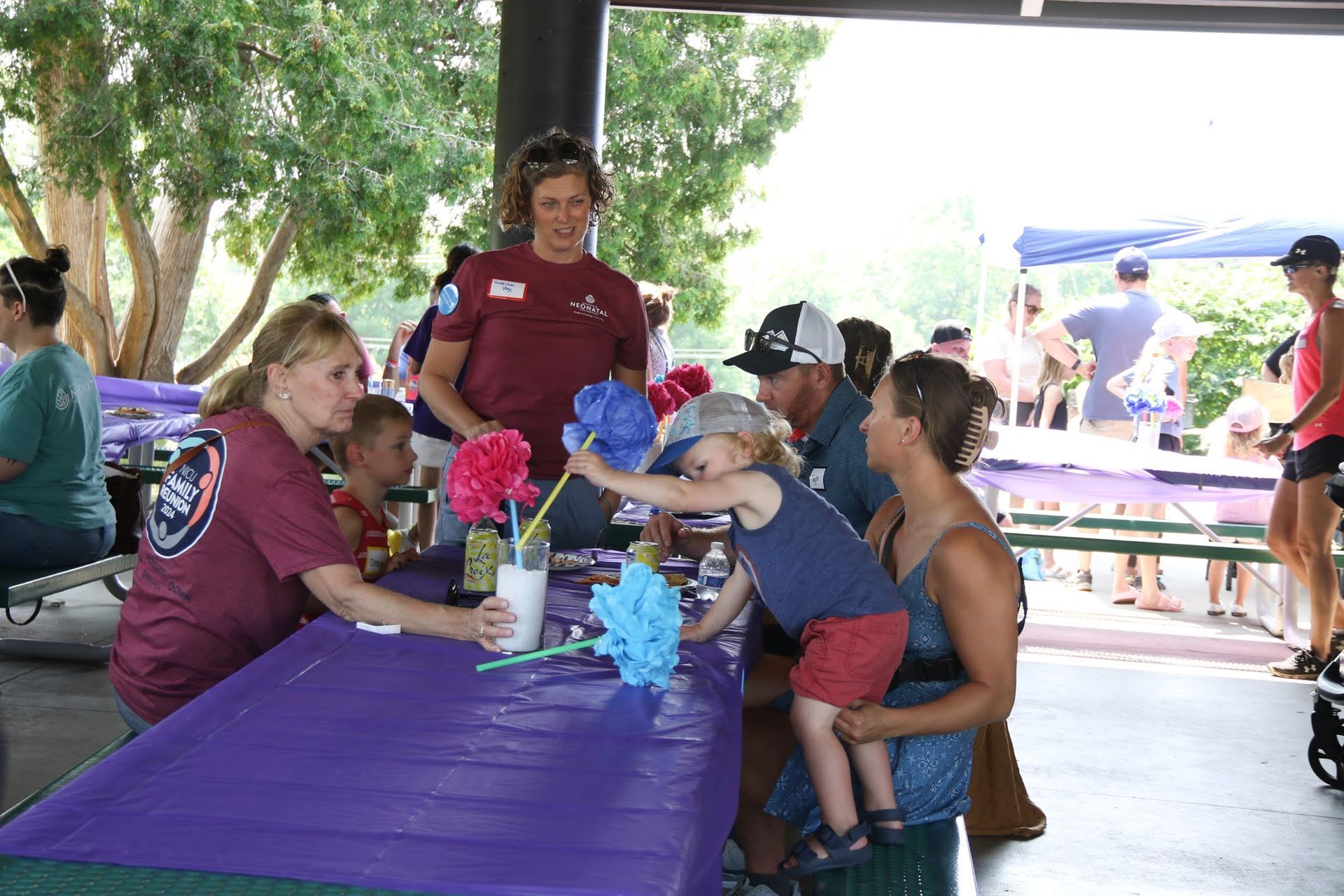 People at a table doing a craft. Child reaches for a flower, being held by parent. Park setting.