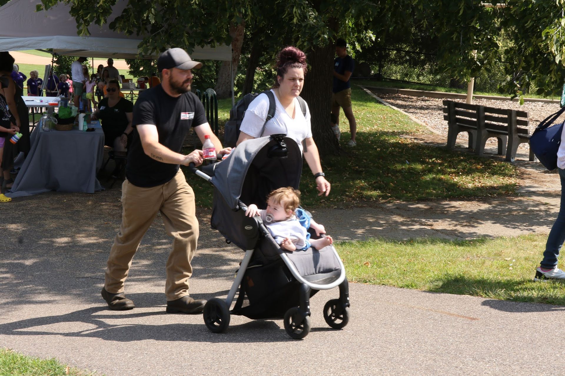 Family pushing a stroller on a paved path in a park; a child is seated in the stroller.