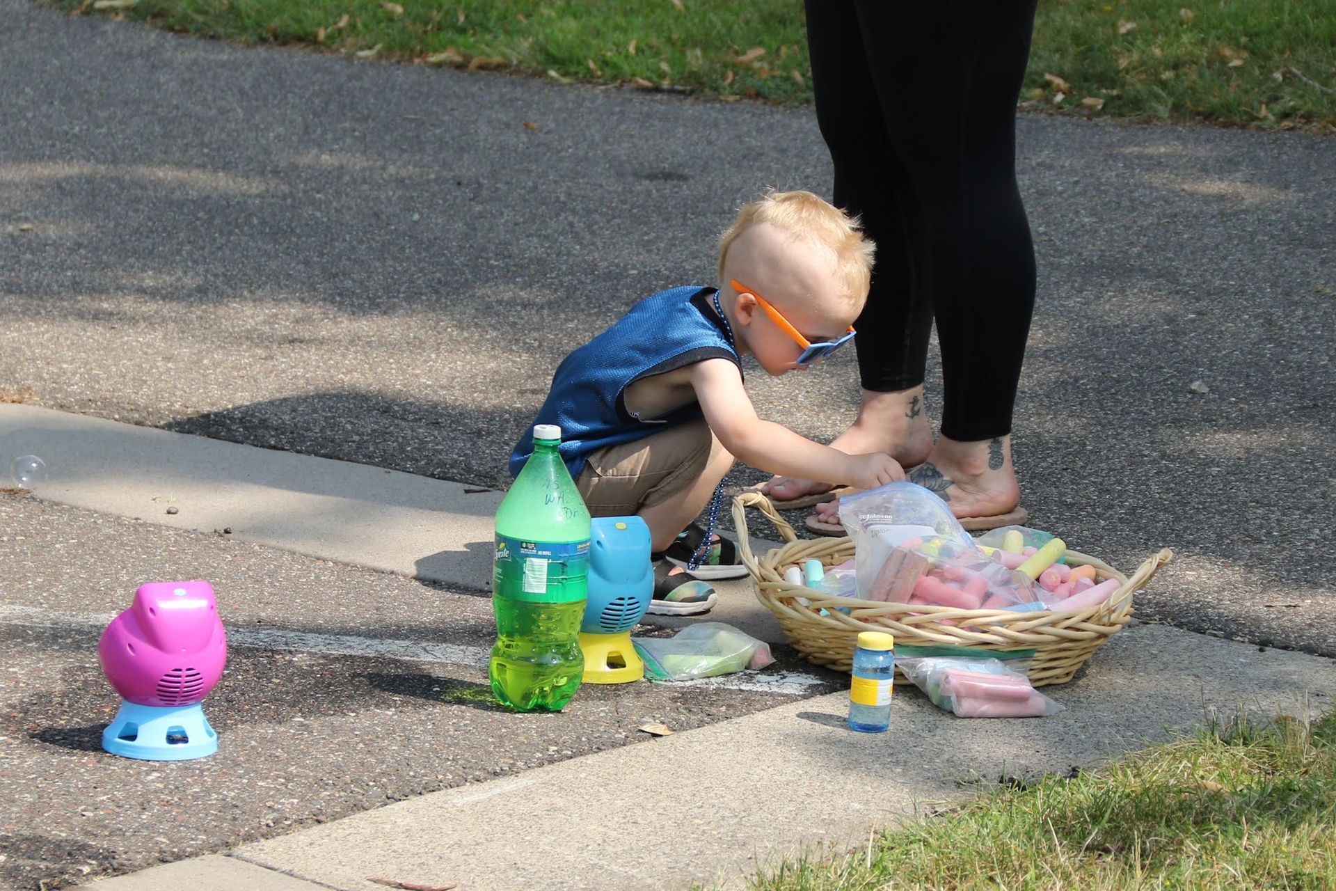 Child reaching into a basket of chalk on a sidewalk, near a soda bottle and a bubble machine. Adult's legs visible.