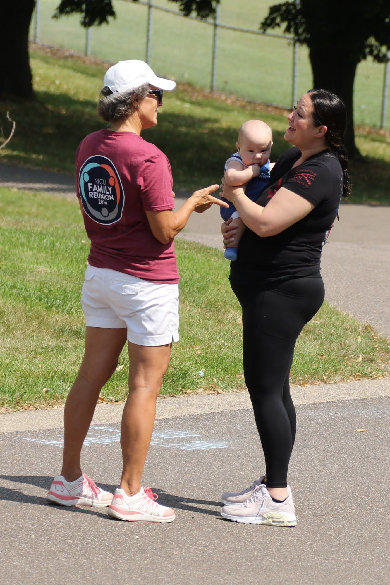 Two women and a baby outdoors; one woman holds the baby, the other points; sunny day.