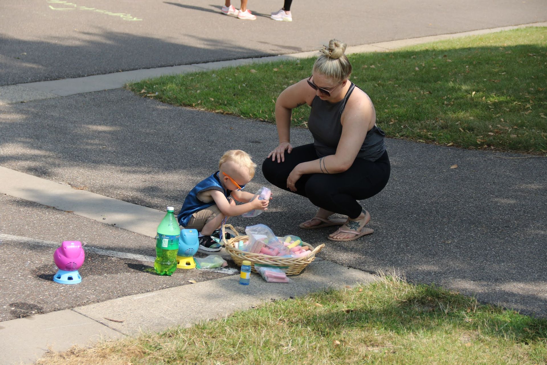 Woman and toddler on pavement, looking at items from a basket. Green grass and sidewalk visible.