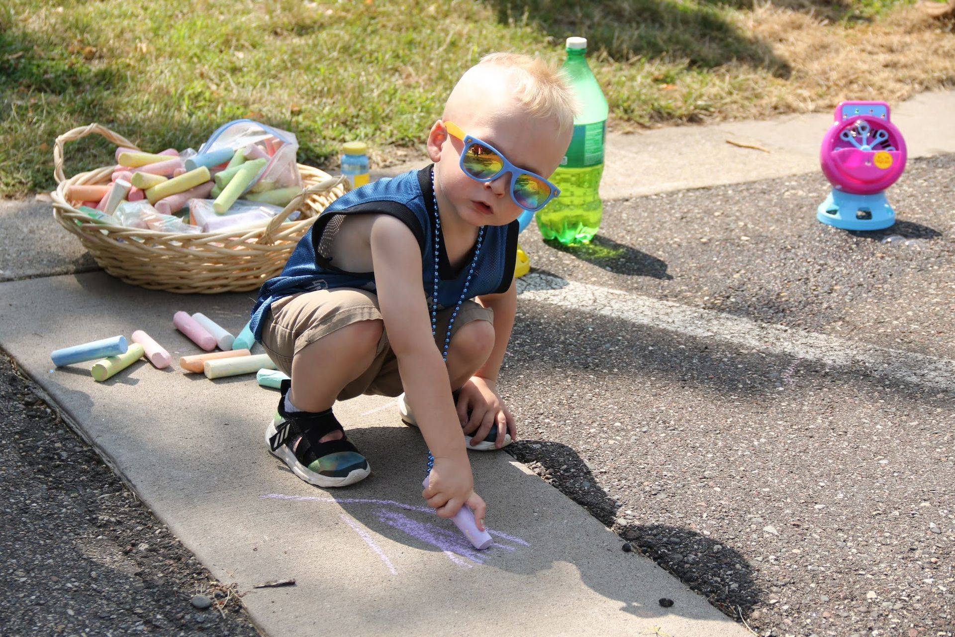 Boy with sunglasses drawing on a sidewalk with chalk. Basket of chalk, water bottle, and toy visible. Sunny outdoor setting.