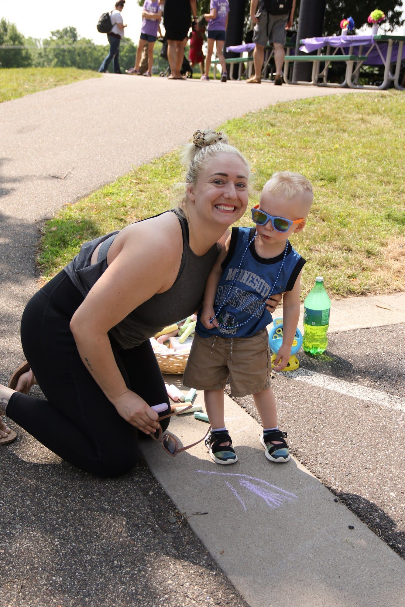 Woman kneels with a toddler, drawing on a sidewalk with chalk at an outdoor event.