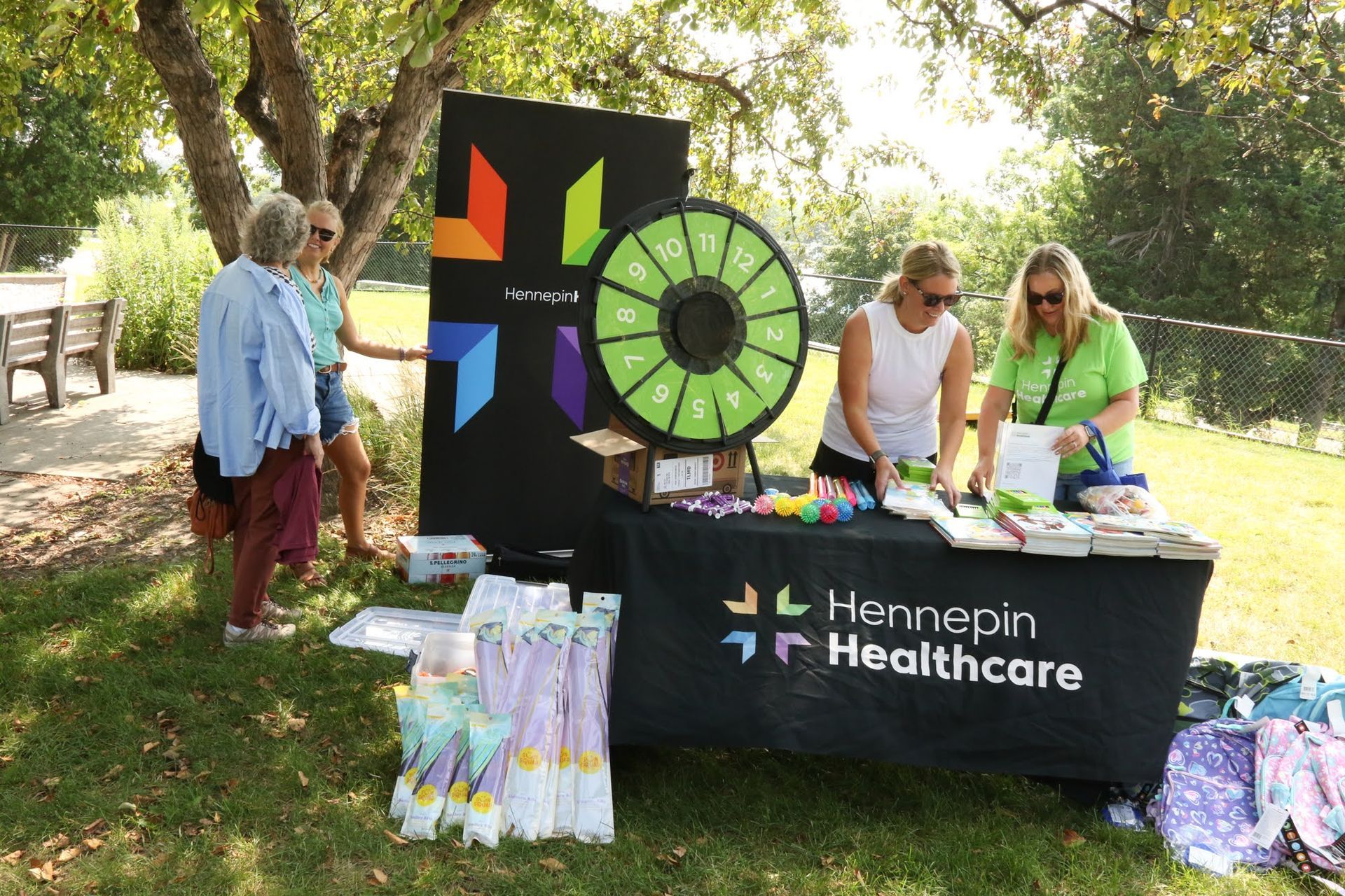 People at a Hennepin Healthcare booth outdoors; spin wheel, table with giveaways, two workers, and two visitors.