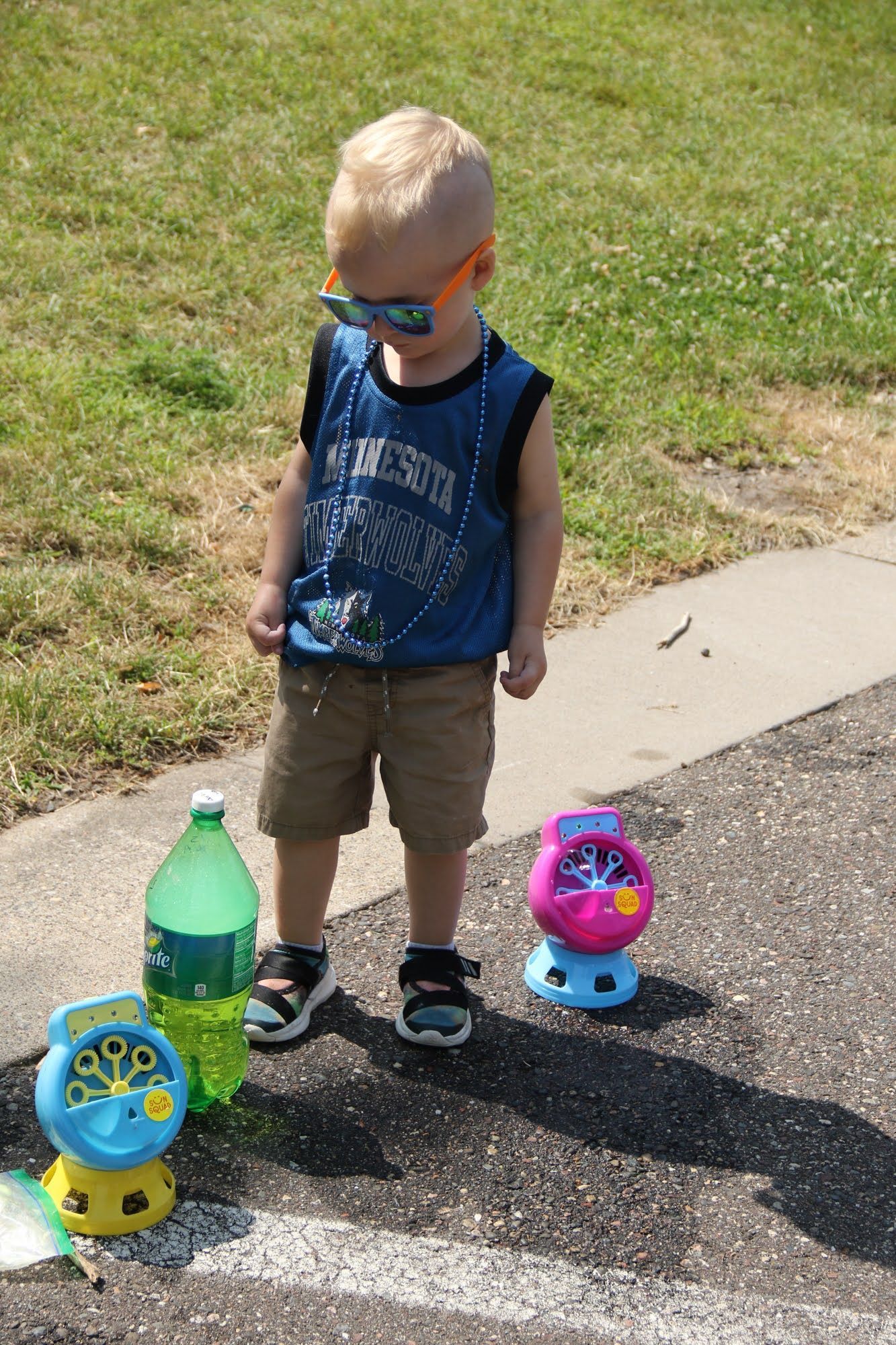 Young child wearing sunglasses, standing near bubble machines and a soda bottle on a paved surface.