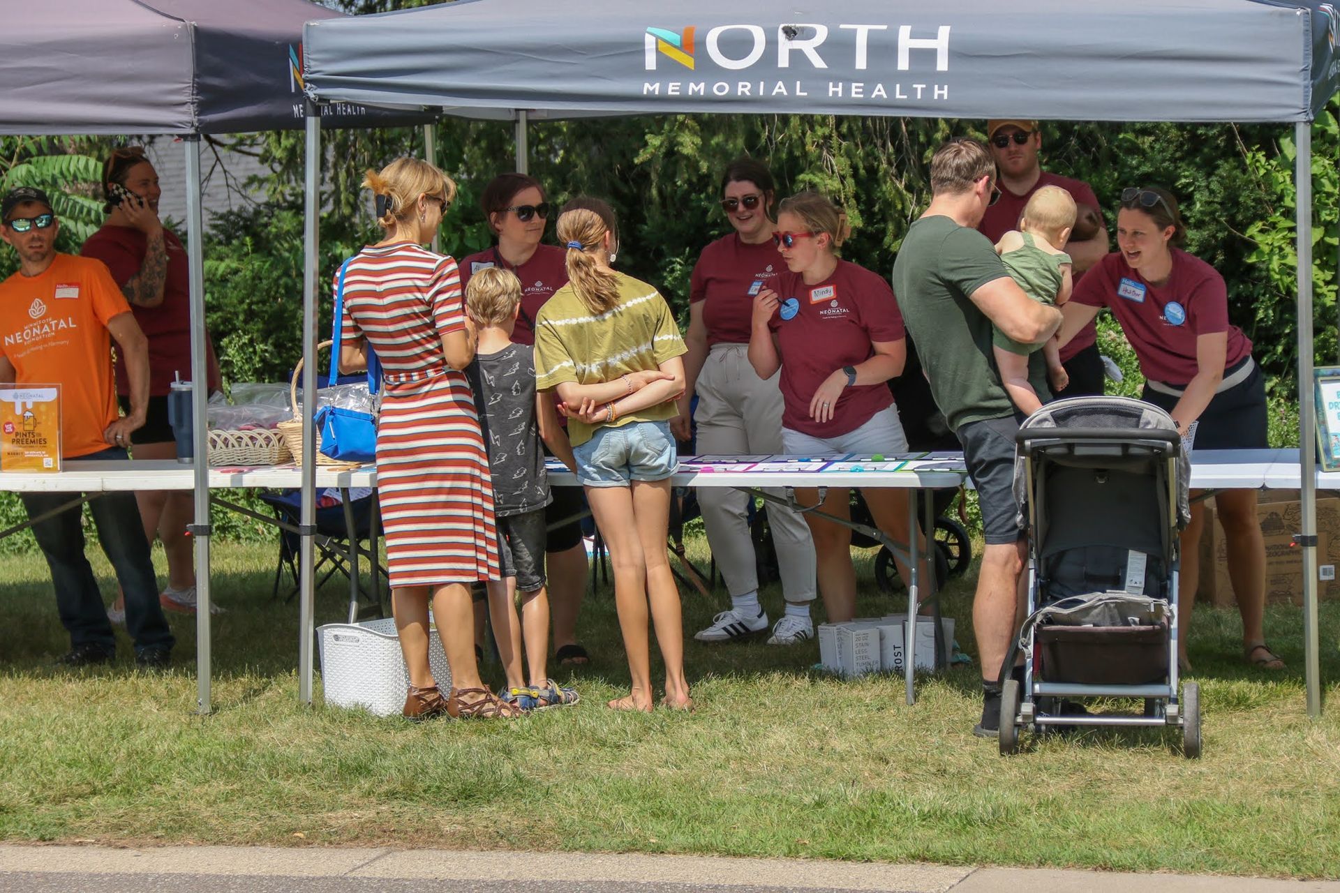 People at a North Memorial Health tent outdoors, some wearing maroon shirts, offering information and items.