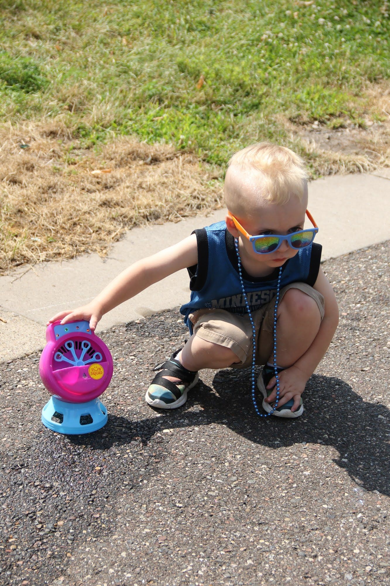 Boy in sunglasses, squatting next to a bubble machine on pavement outdoors.