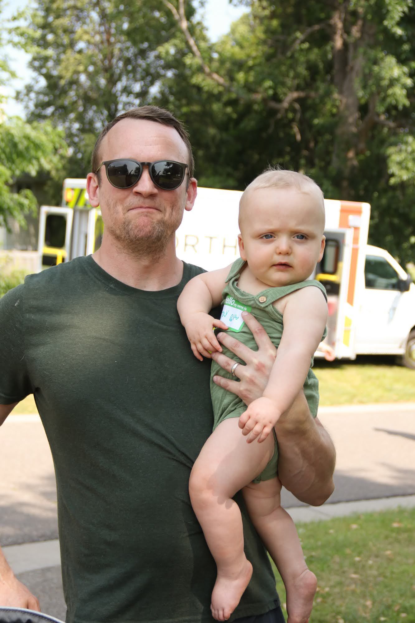 Man in sunglasses holds baby in a green onesie. Both looking at the camera. Outdoor setting with a white vehicle.