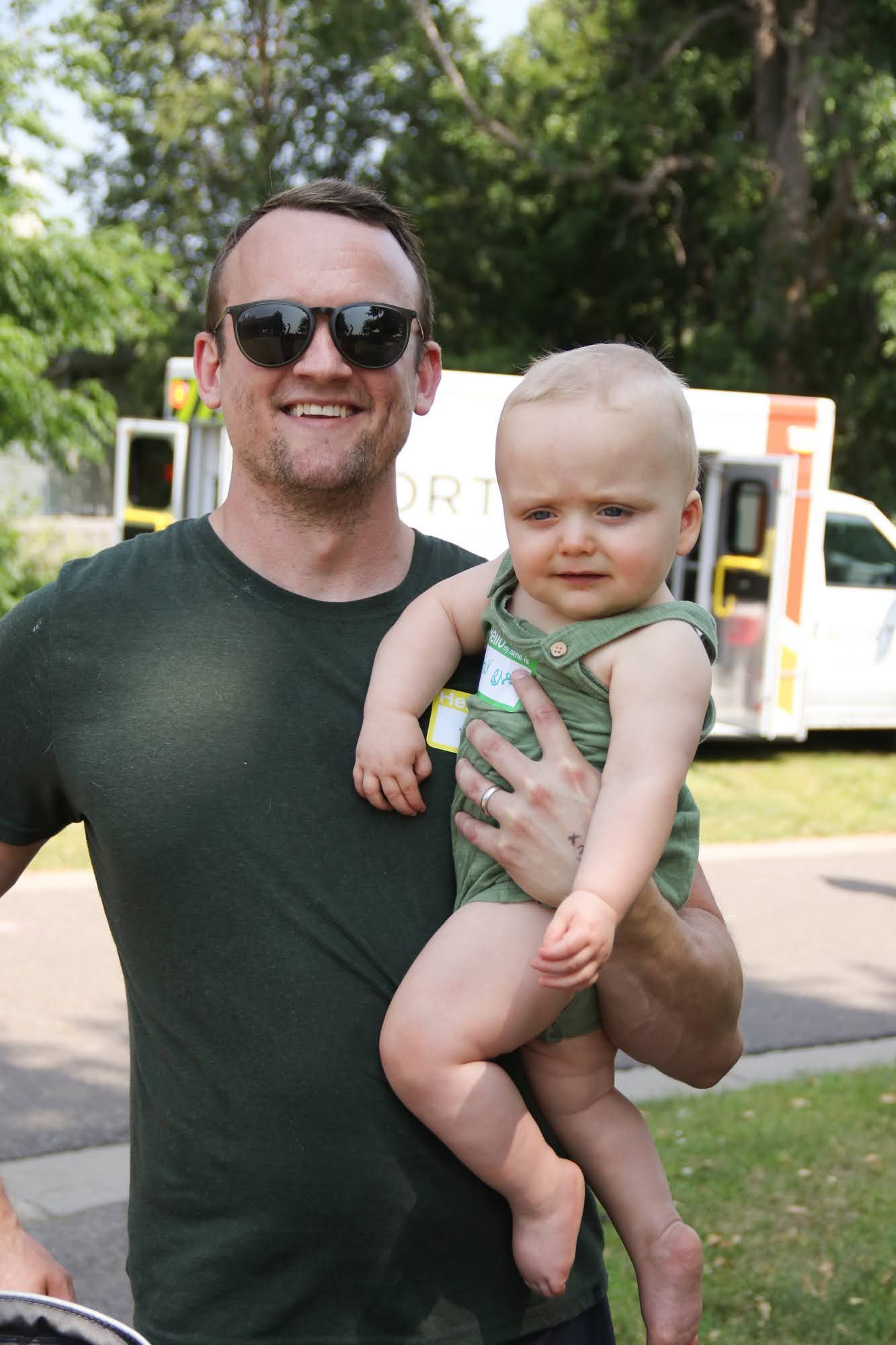 Man in sunglasses holds a baby in green onesie outside, with an ambulance in the background.