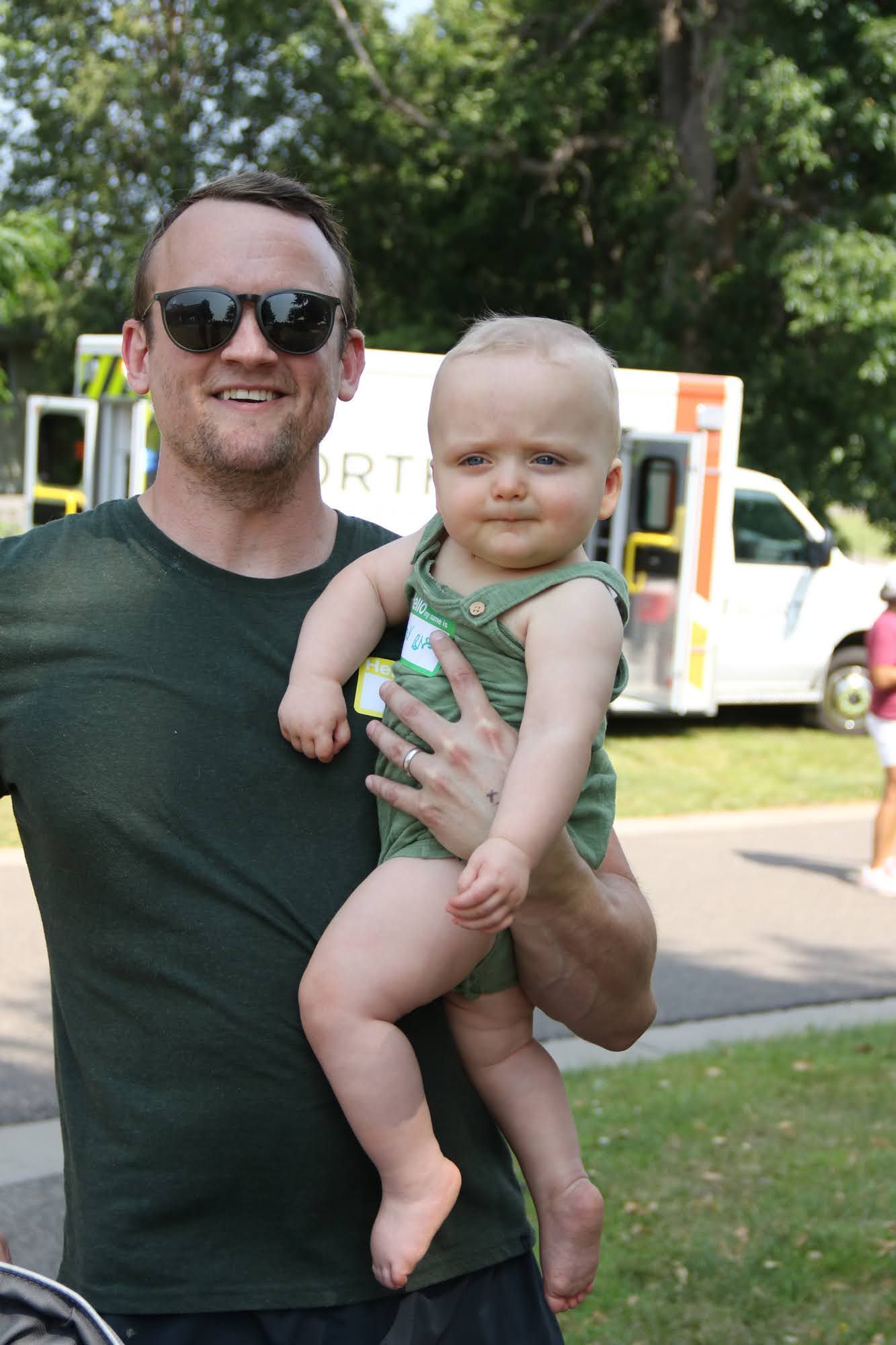 Man in sunglasses holding a baby in a green onesie; parked ambulance in the background.