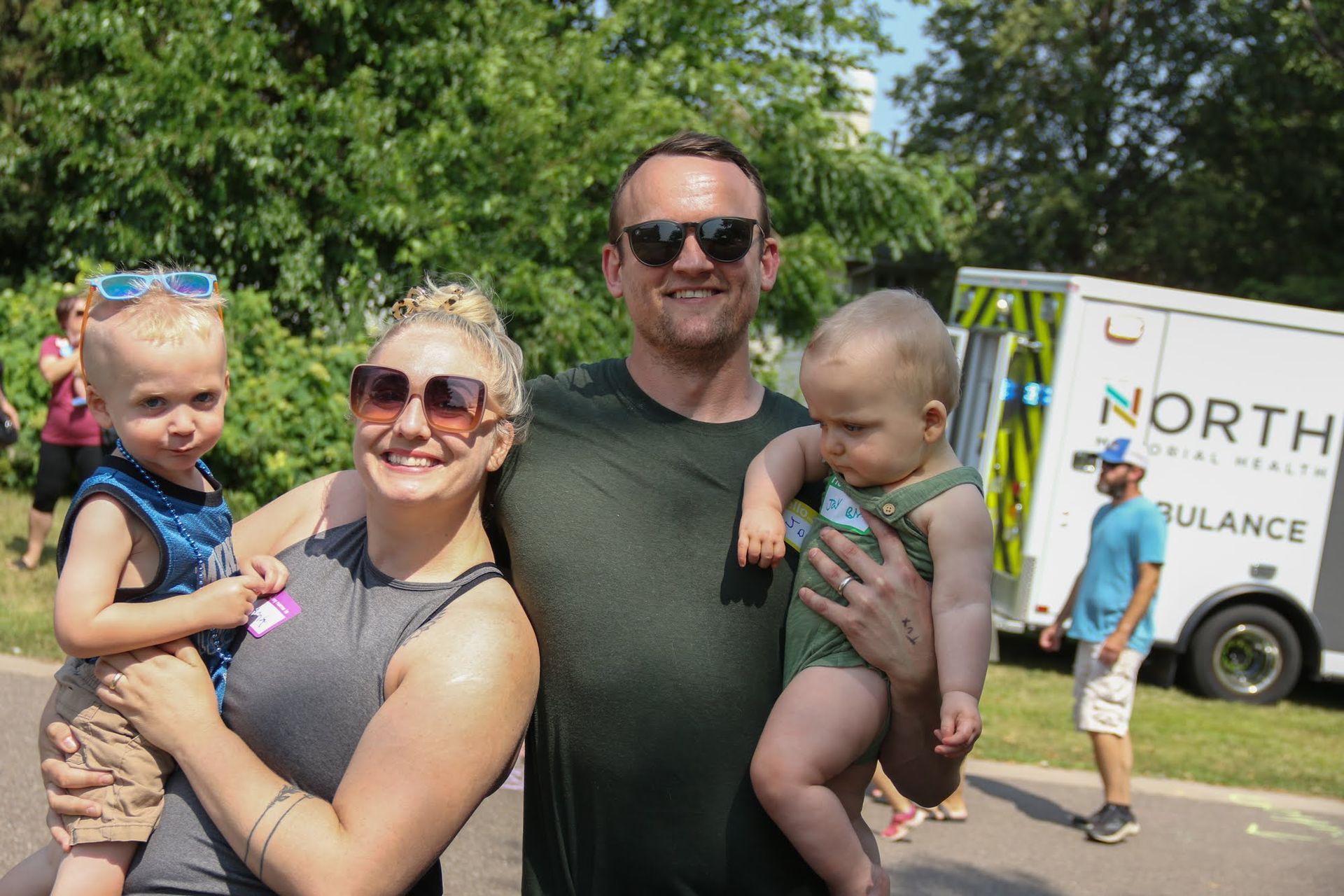 Family smiling outdoors, holding children; ambulance in background.