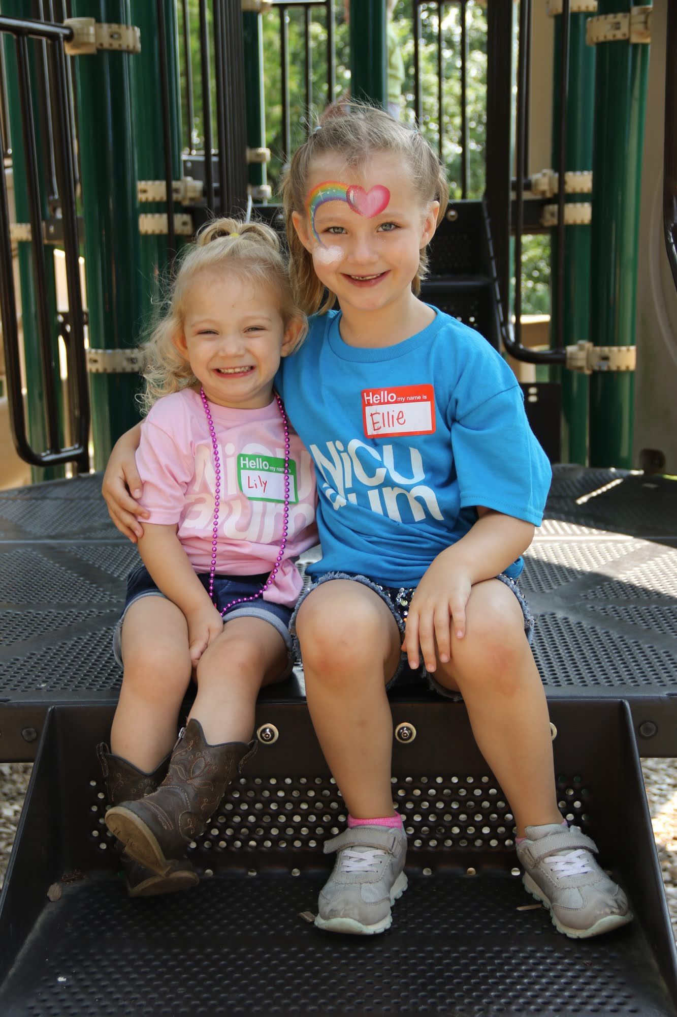 Two young girls smiling and sitting on playground steps. One has face paint and a blue shirt, the other wears a pink shirt and cowboy boots.