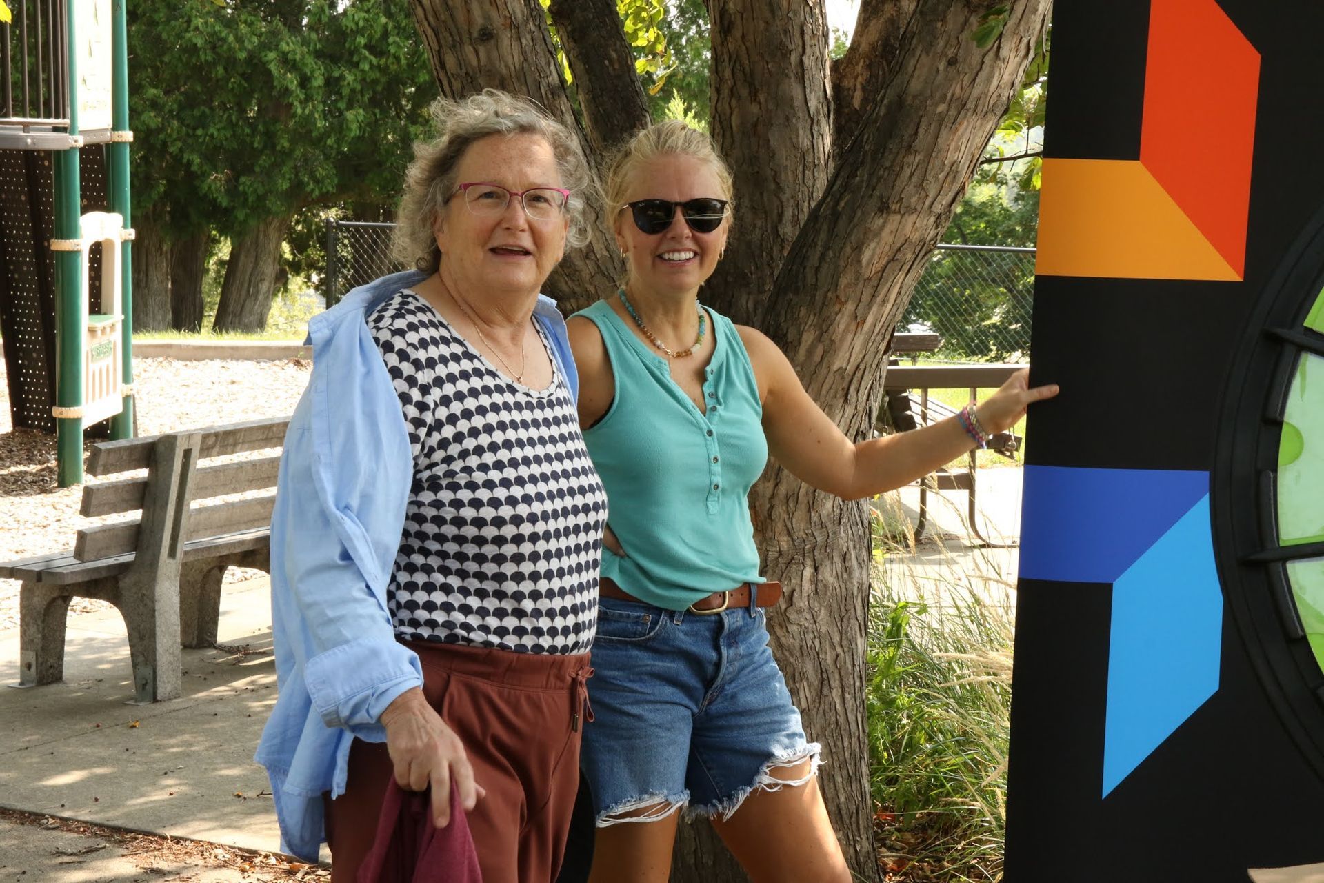 Two women pose beside a colorful abstract mural in a park. One points to it, smiling.