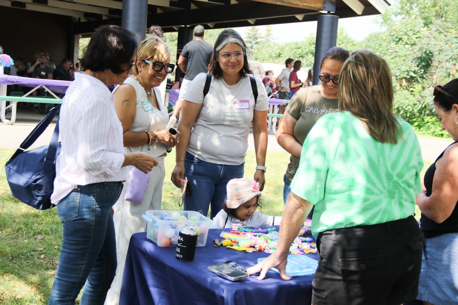 People gather at an outdoor table with a child, possibly at a community event.