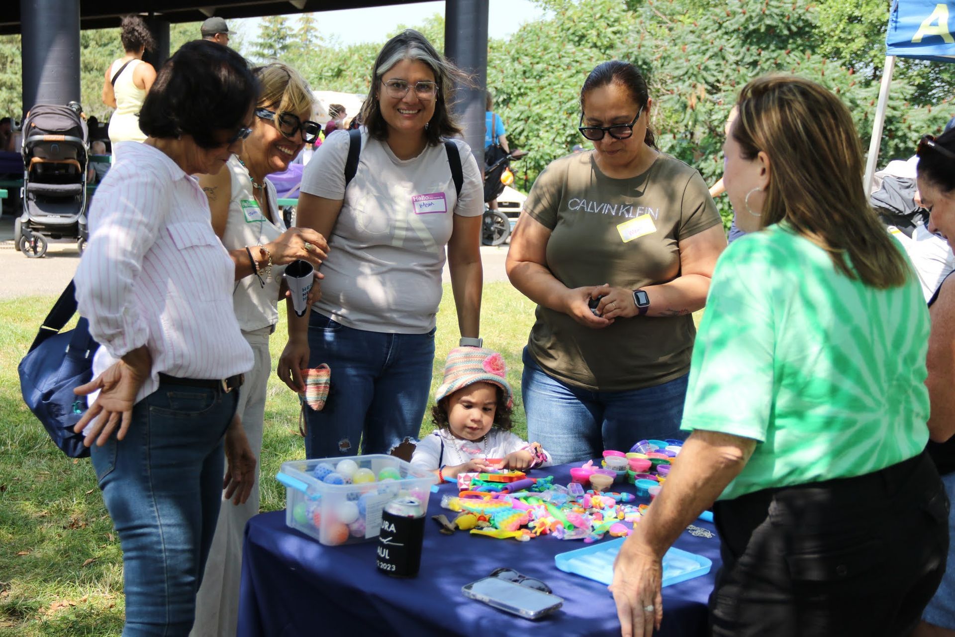 People gathered around a table with items, a child sits in front. Outdoors in sunlight.