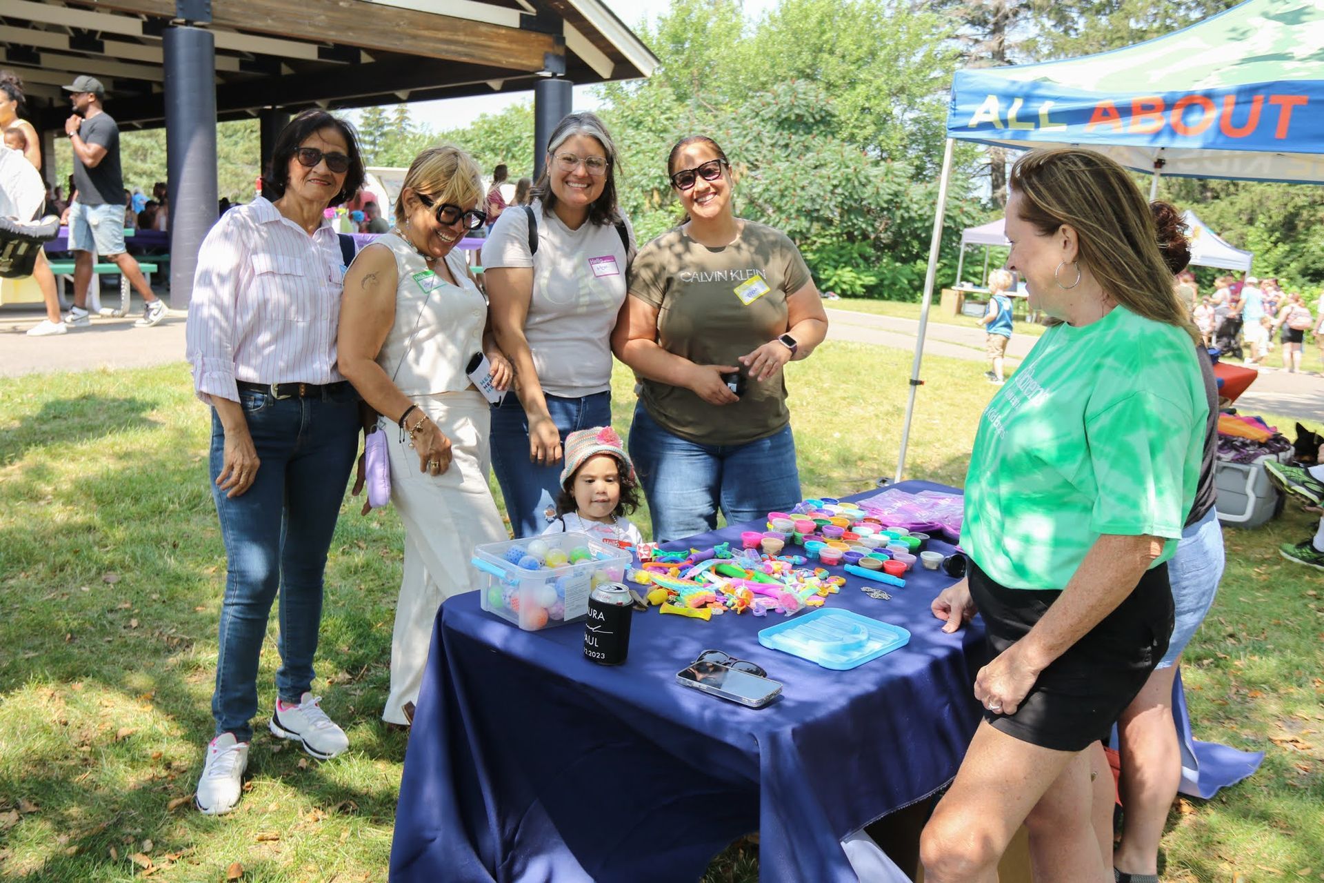People at a table with colorful items outside. One woman wears a green shirt.