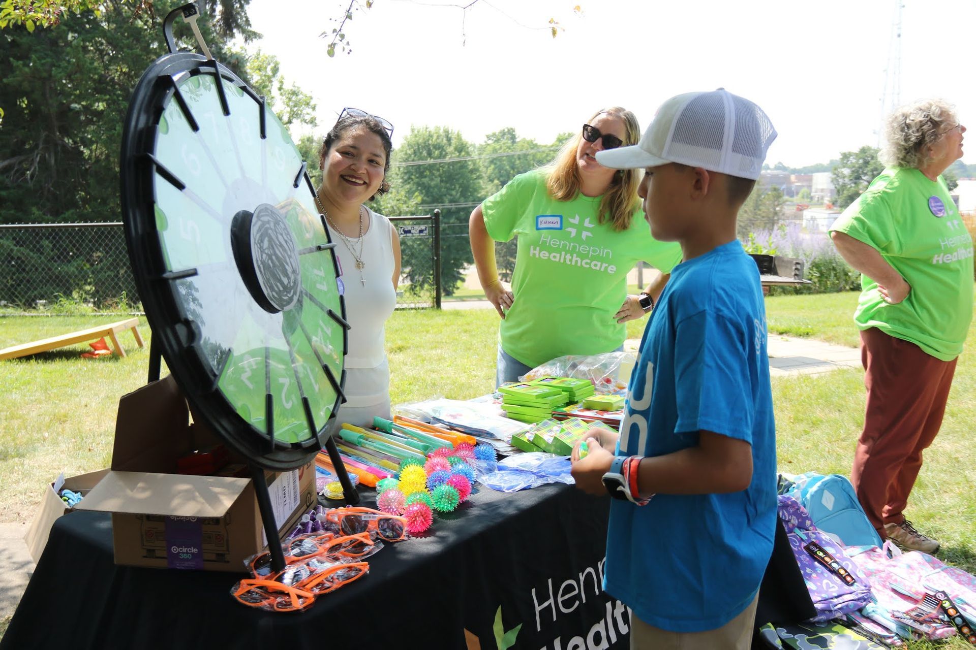 A boy spins a prize wheel at an outdoor event table with two women.