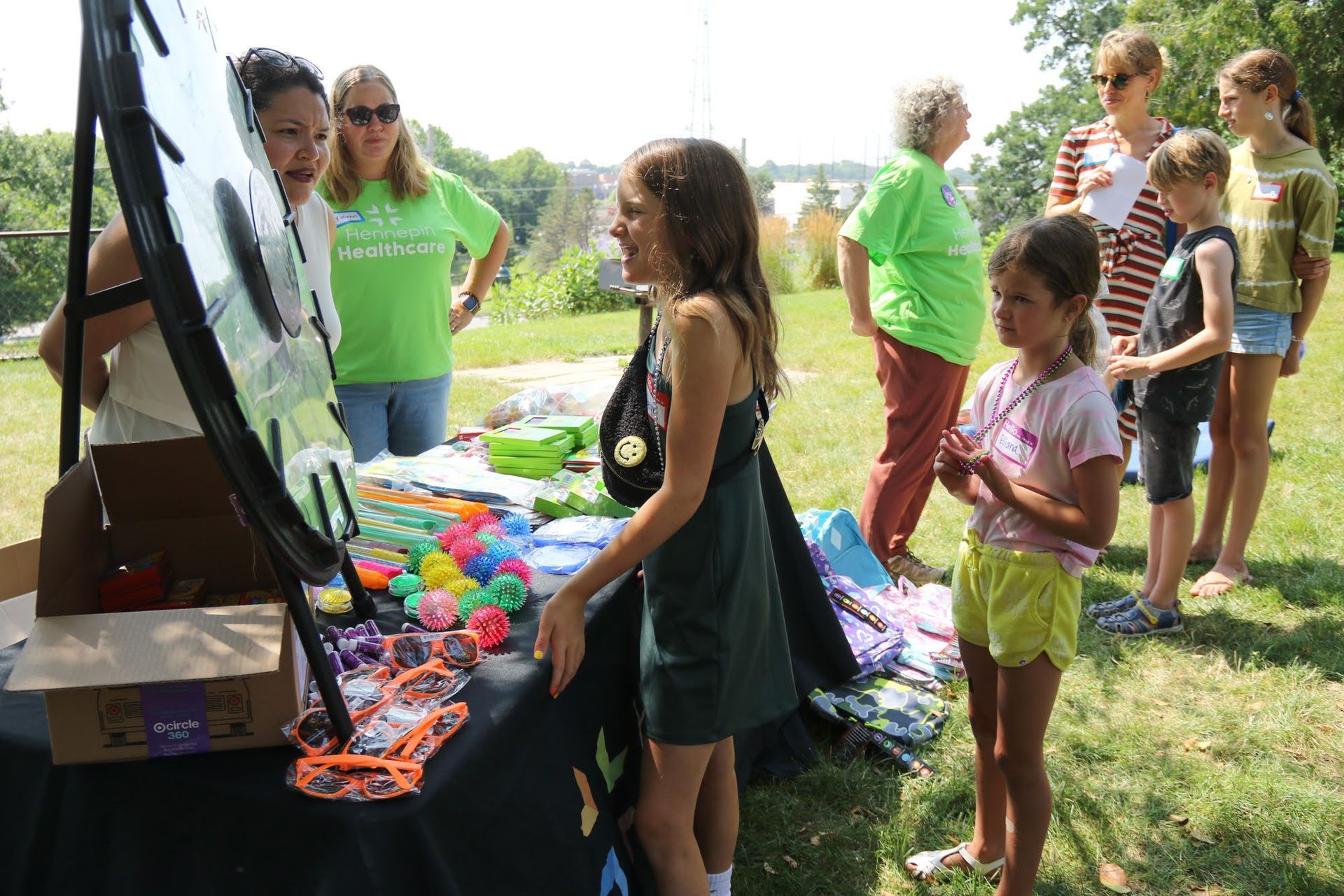 People at an outdoor event with a table of toys. Girl smiles as she looks at the items. Green grass and trees are in the background.