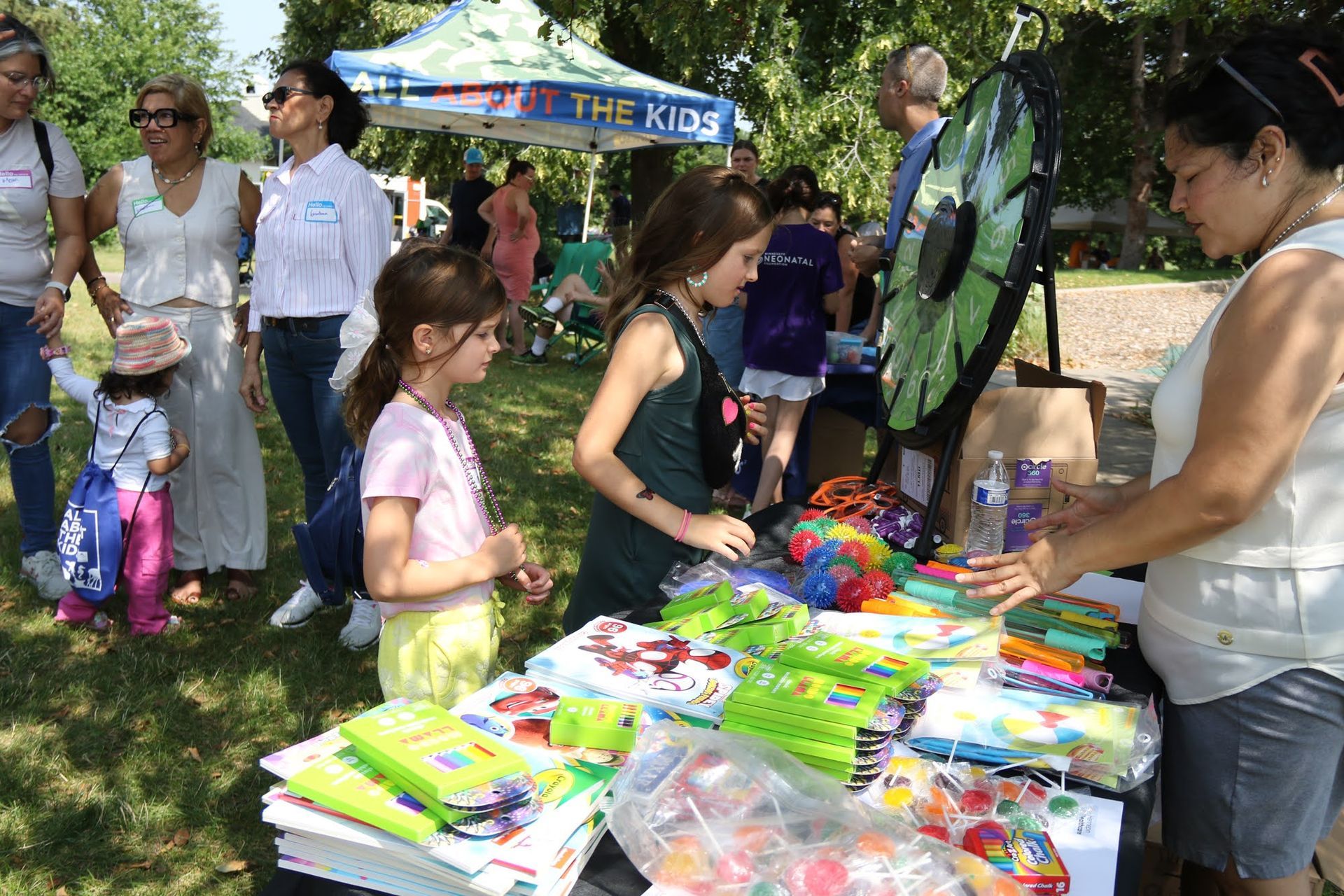 People at a table with school supplies and a prize wheel; outdoors, sunny day.