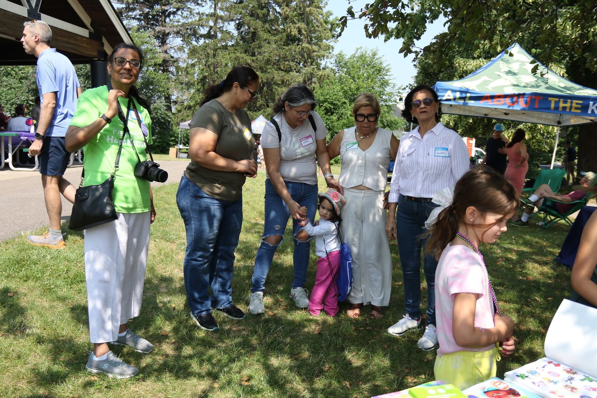 People gather outdoors near a pavilion and a tent. One woman in green smiles, others chat.