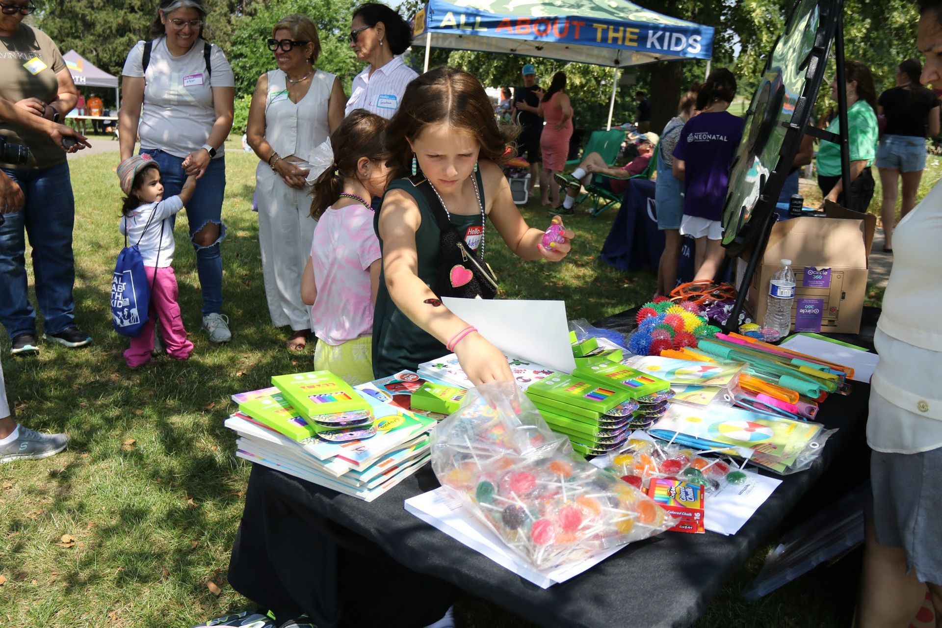 Girl at a craft table selling items at an outdoor event, with people nearby.