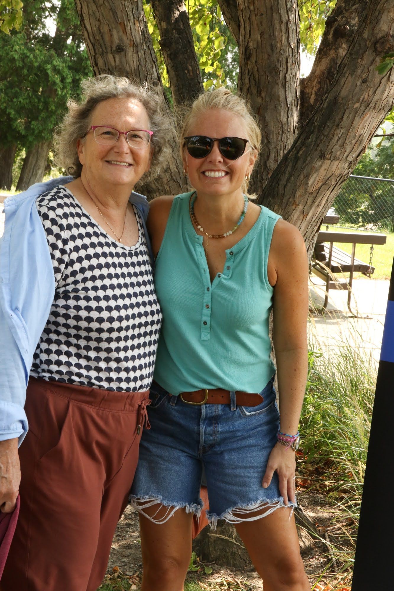 Two women smiling, posing for a photo outdoors near a tree. One wears a checkered shirt, the other a teal top and jean shorts.