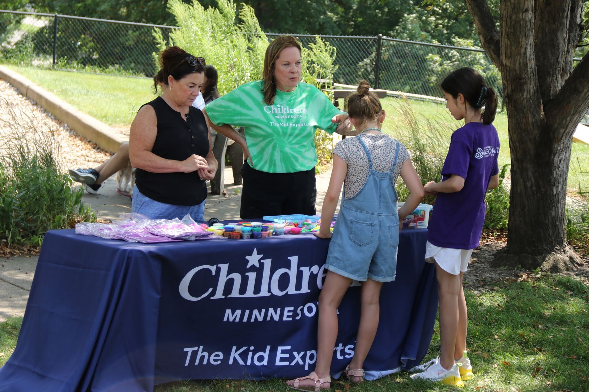 People at Children's Minnesota booth. Two children at a table with staff, blue table, green grass, and trees.