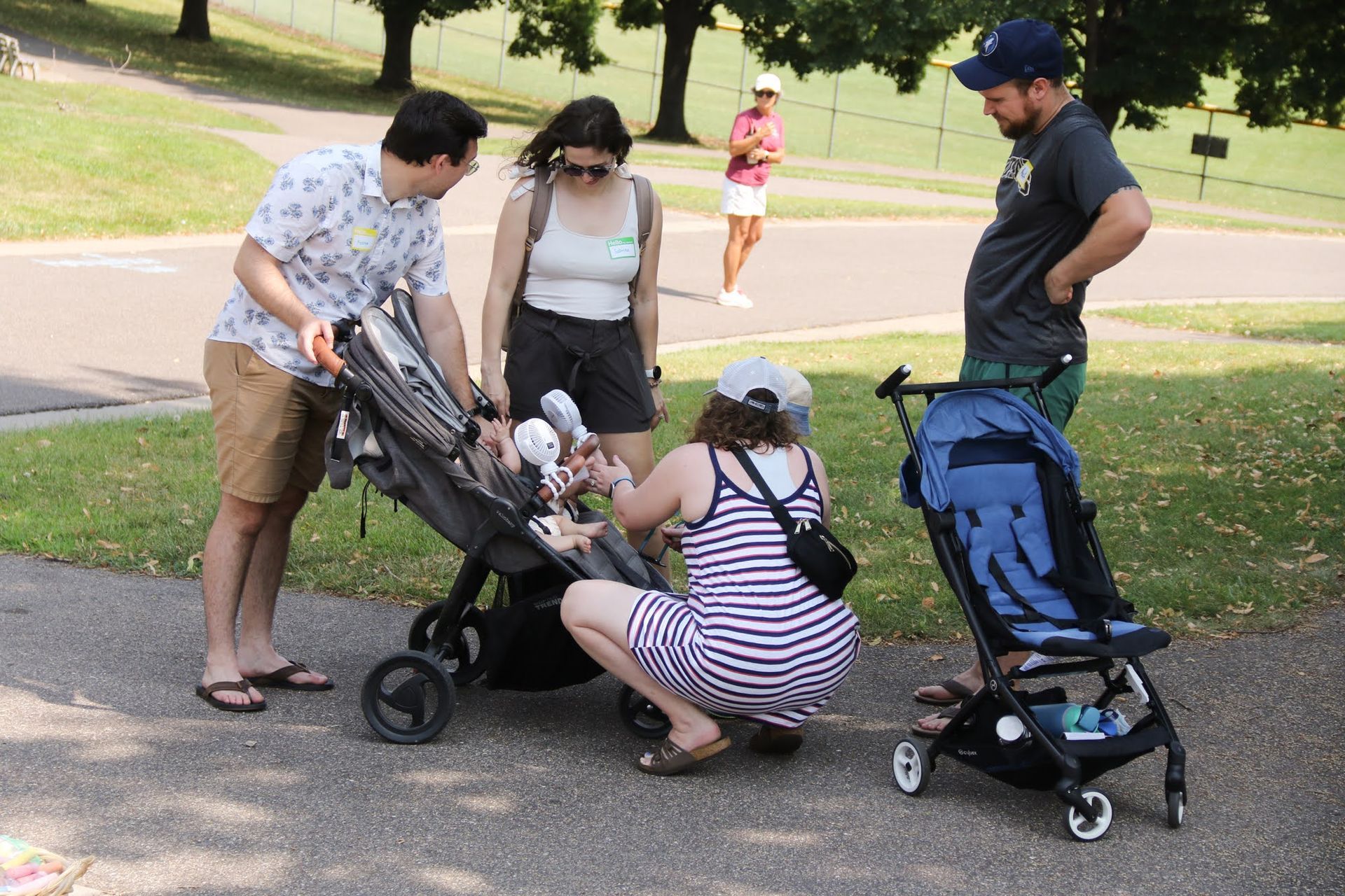 People with strollers on a path in a park; one person kneeling at a stroller, others look on.