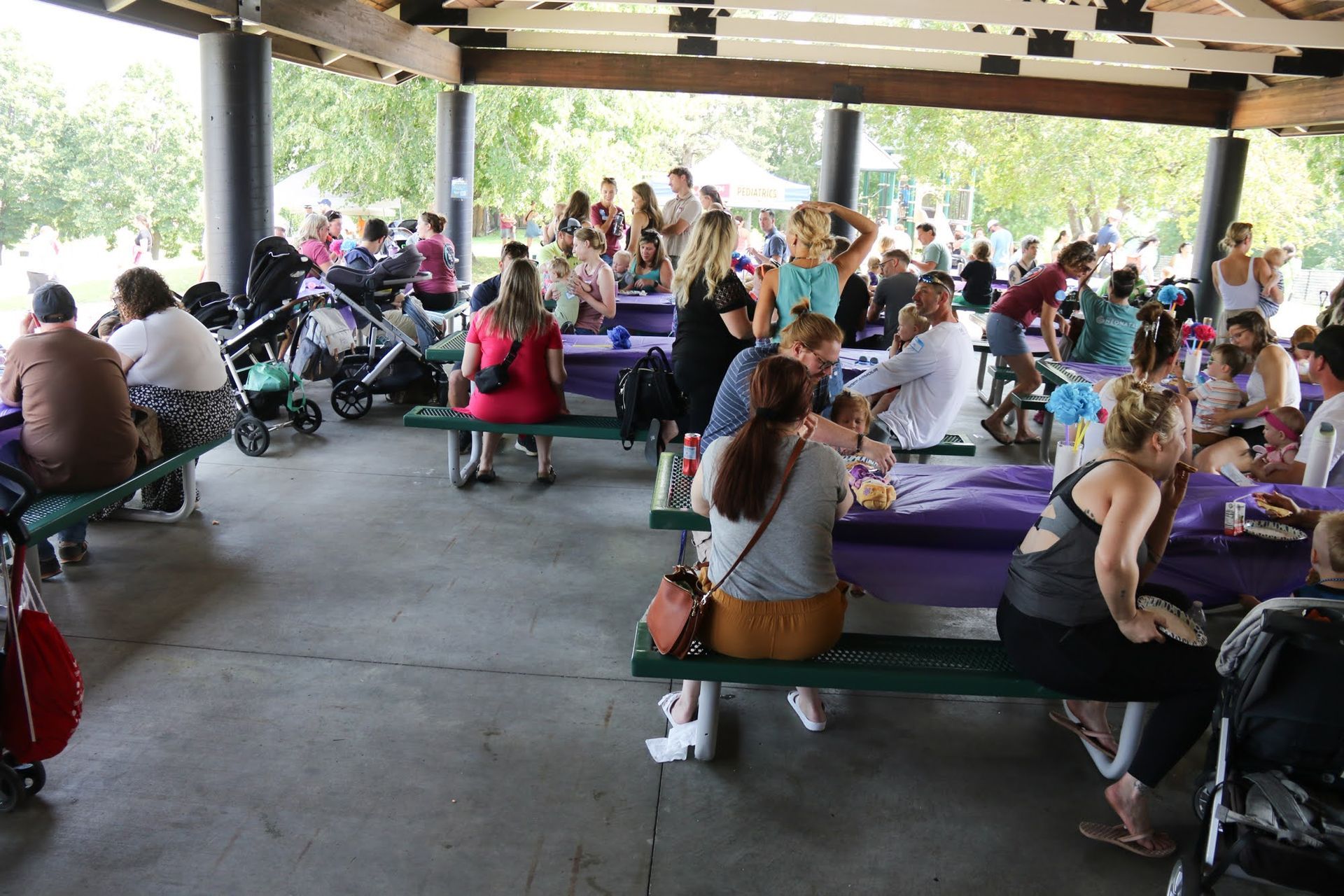 Picnic under a pavilion. Many people at tables, with children, food, and strollers.