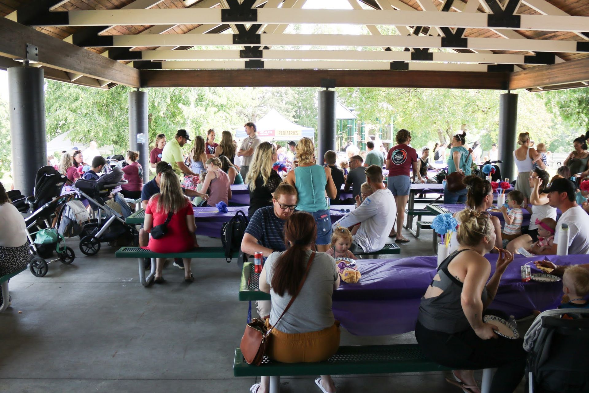 People at picnic tables under a covered shelter in a park.