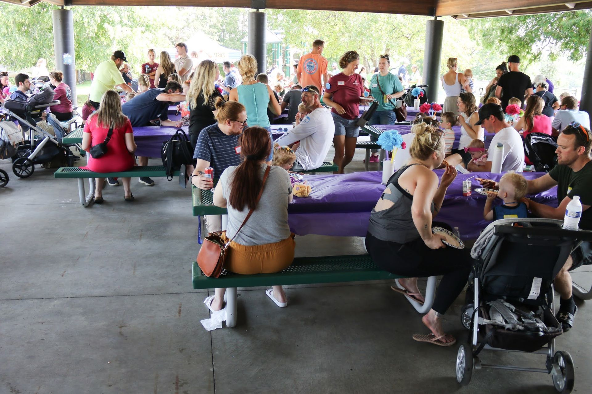 People at a park picnic under a pavilion. Tables with purple tablecloths; strollers.